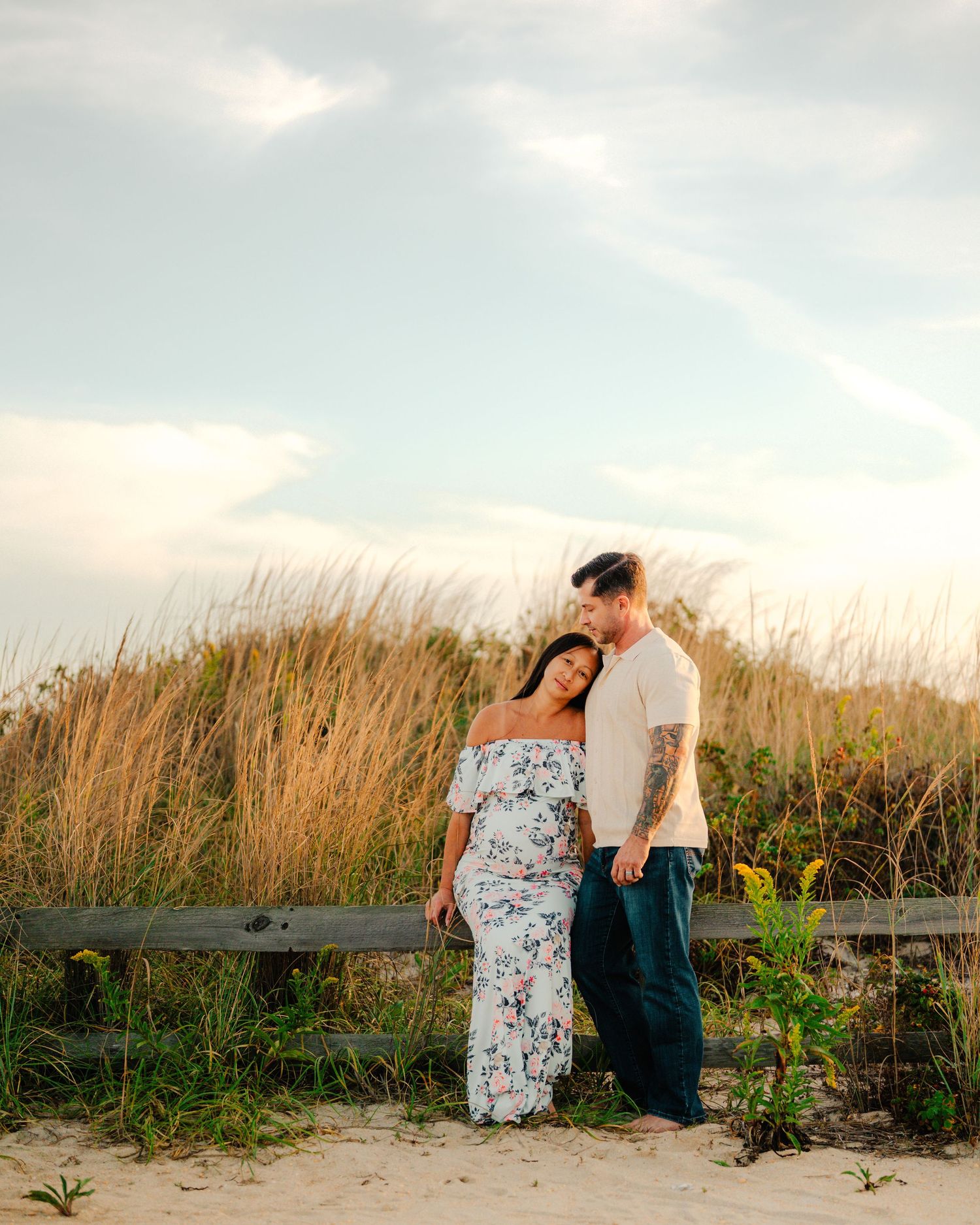 A couple shares a romantic moment on a wooden bench surrounded by tall beach grass at sunset.