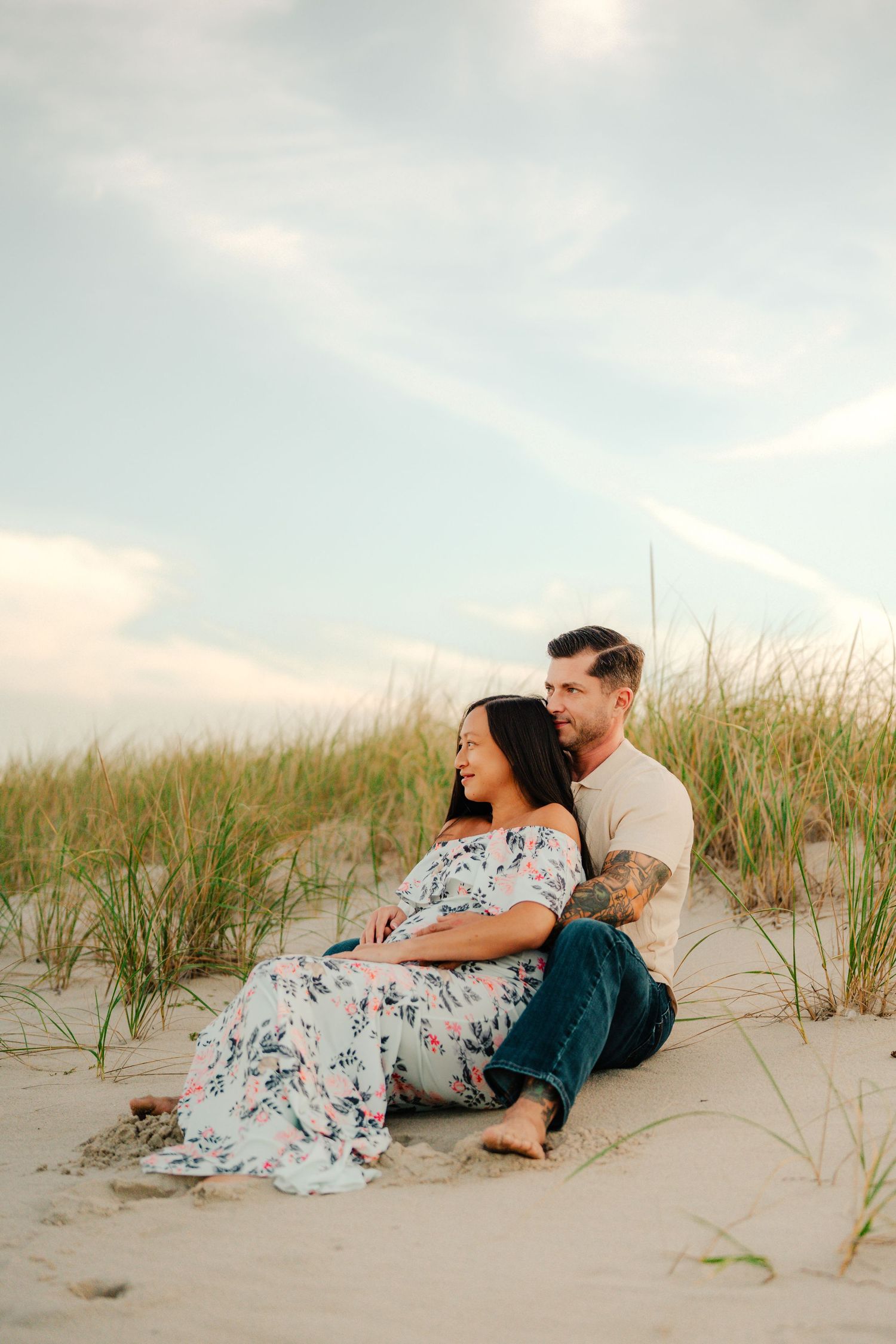 A couple relaxes together on a sandy beach dune with tall grass at sunset.