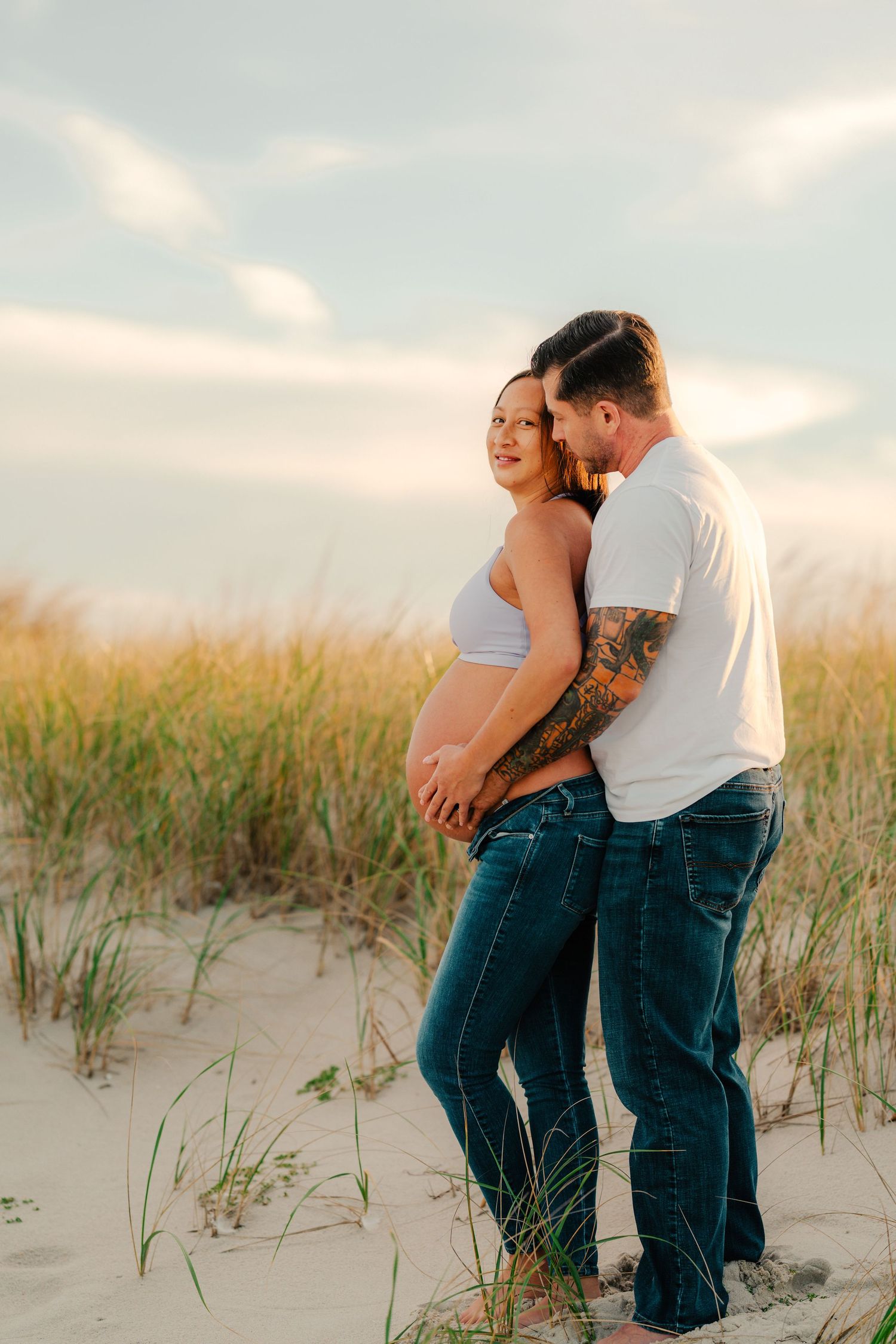 Expectant couple embraces on sandy beach dunes during golden hour sunset with tall grass in the foreground.