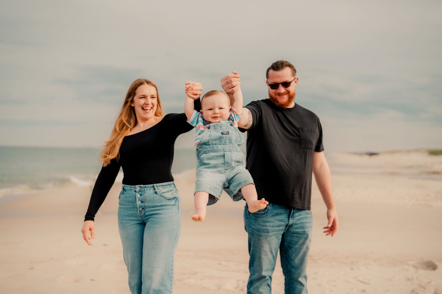 Family in denim and black clothing walking together on sunny beach, swinging child between them while smiling.