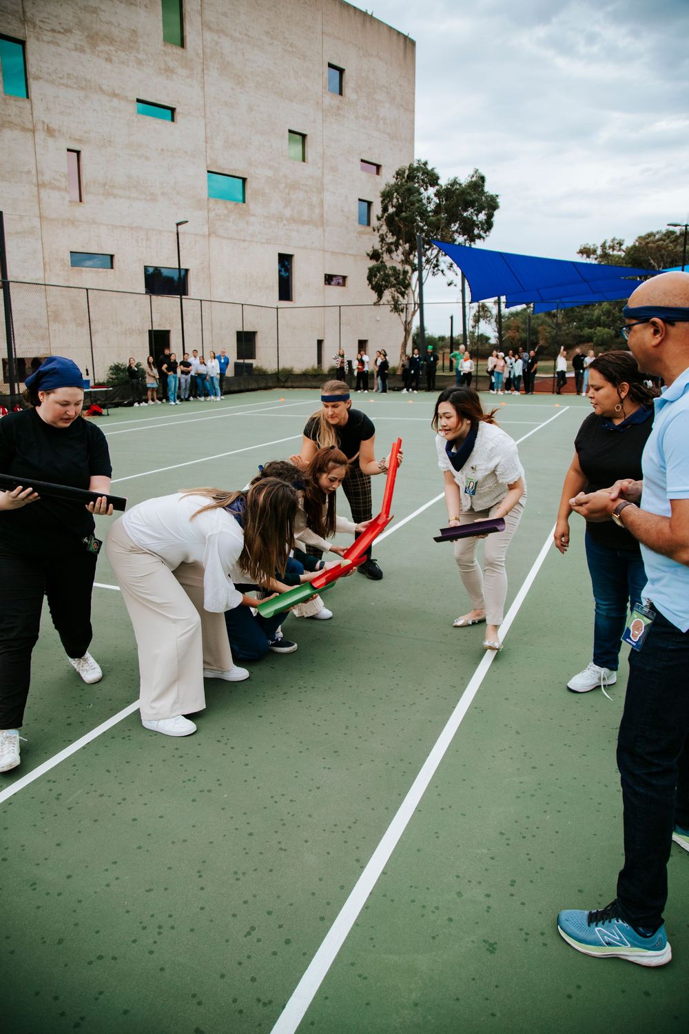 A group of students playing a limbo game under a colorful stick on an outdoor court.