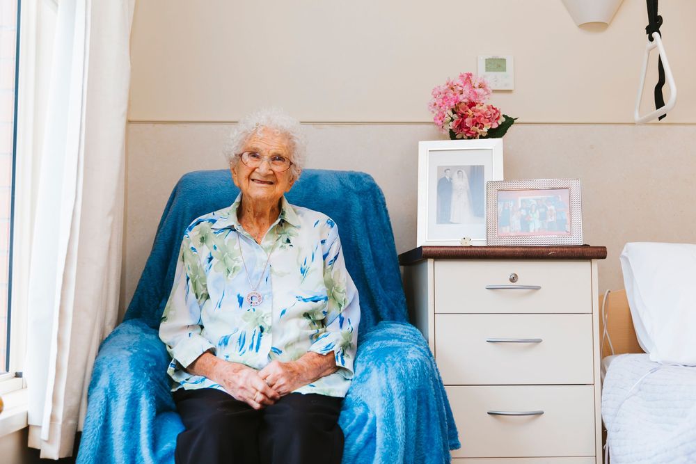 An elderly person sits comfortably in a blue armchair while reading a book in a bright room.