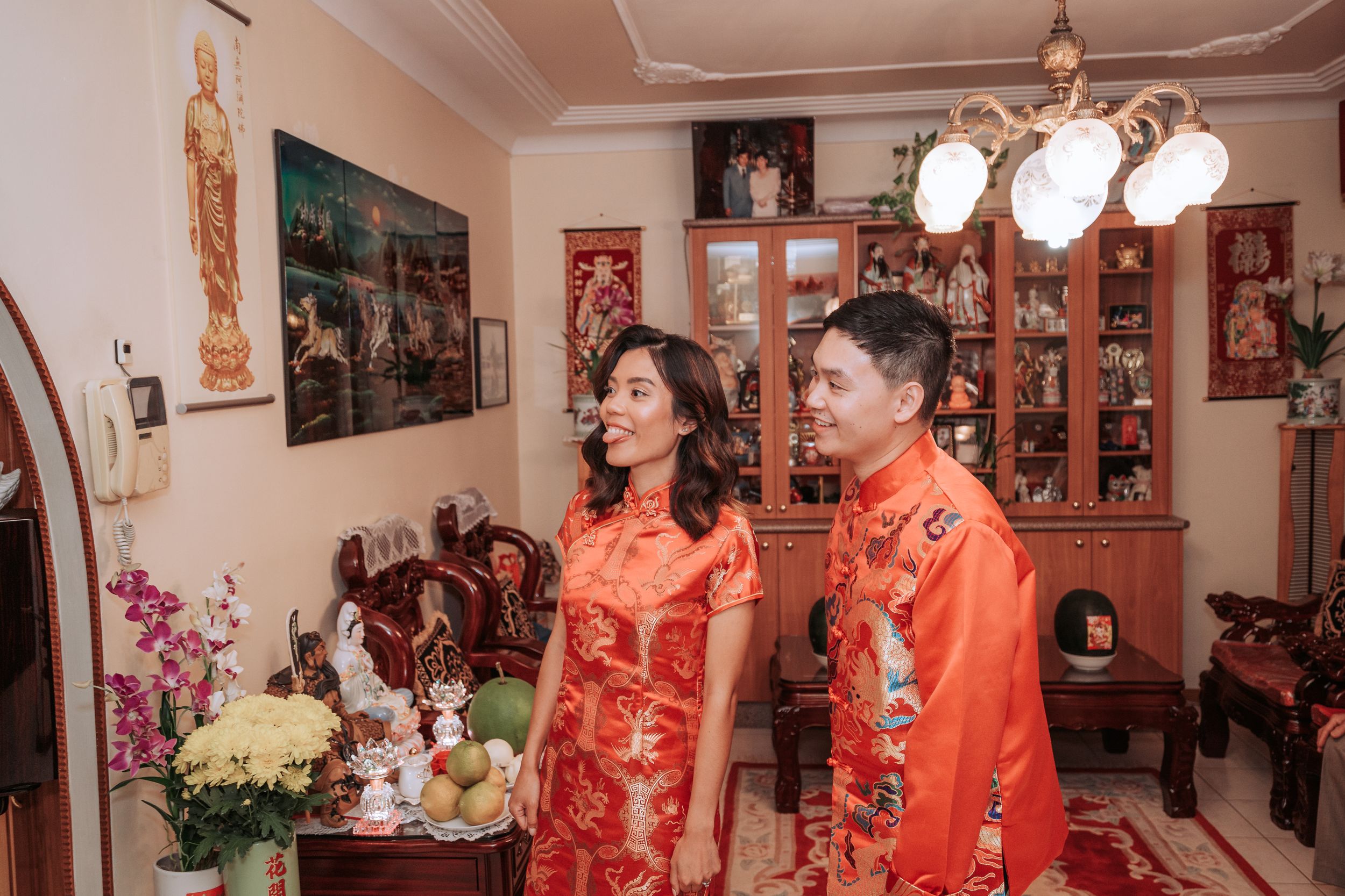 A couple in traditional red Chinese wedding attire posing in their decorated home.