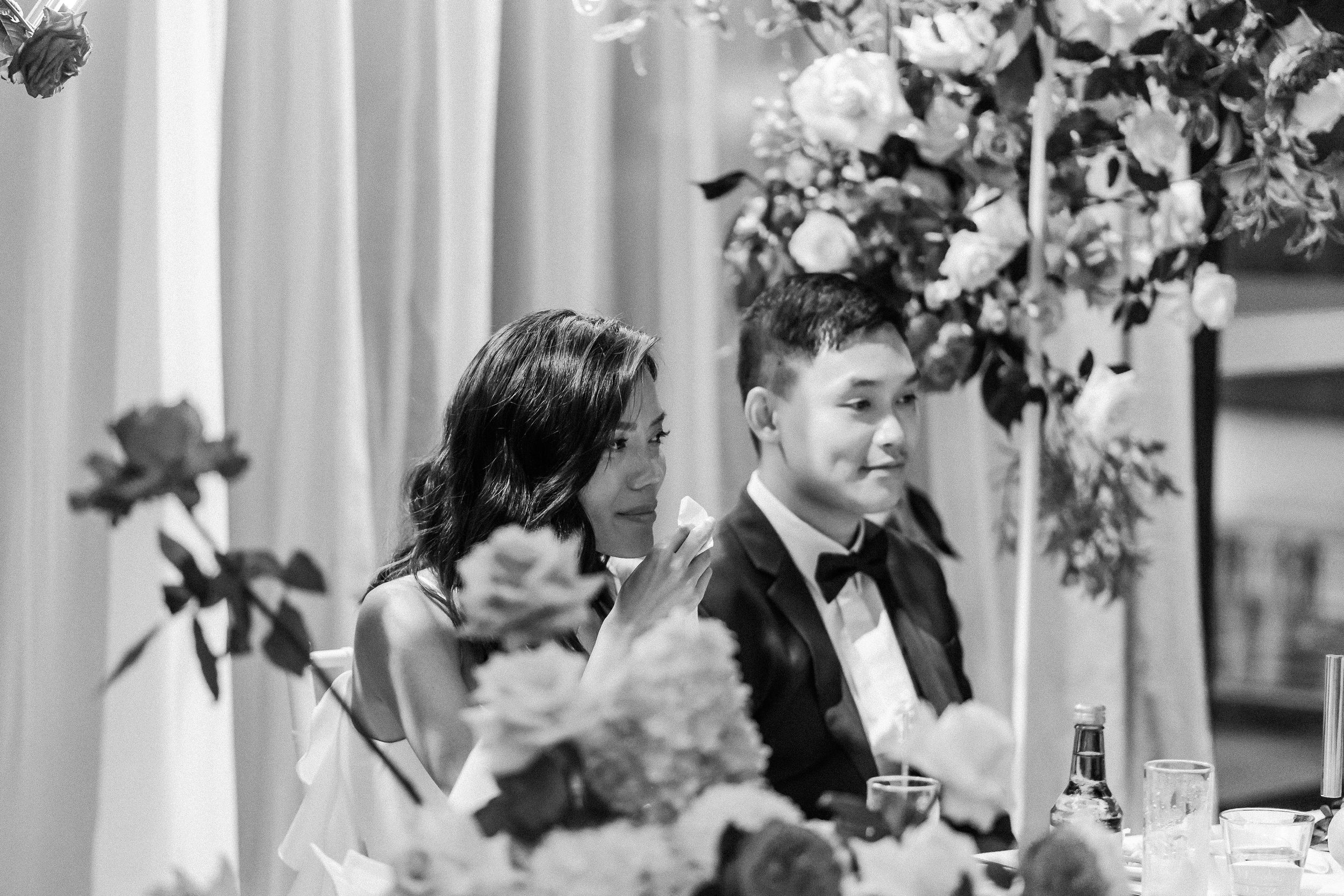 Elegant black and white photo of newlyweds at their reception dinner table surrounded by floral arrangements.