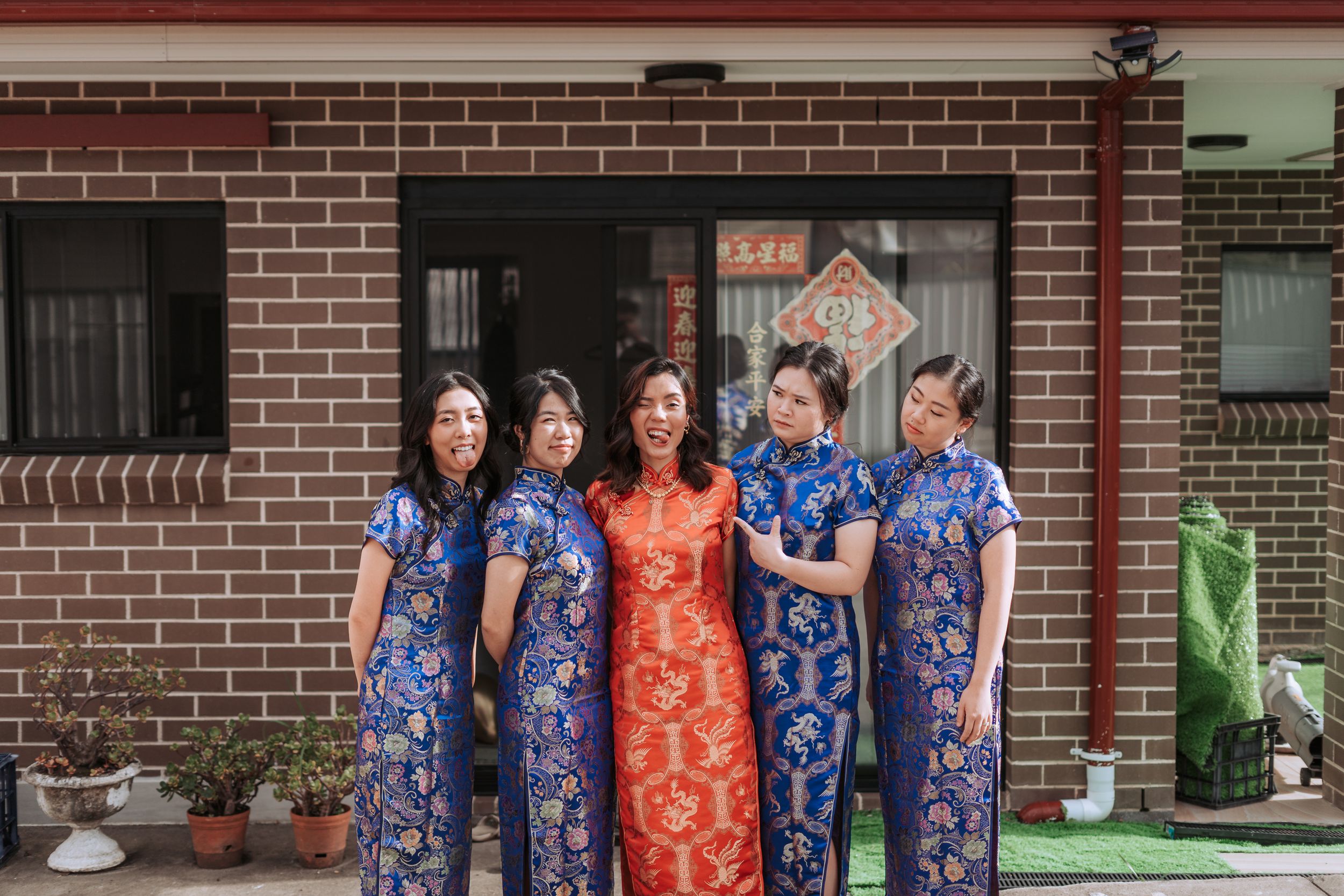 Five women in traditional Chinese qipao dresses posing in front of a brick house.