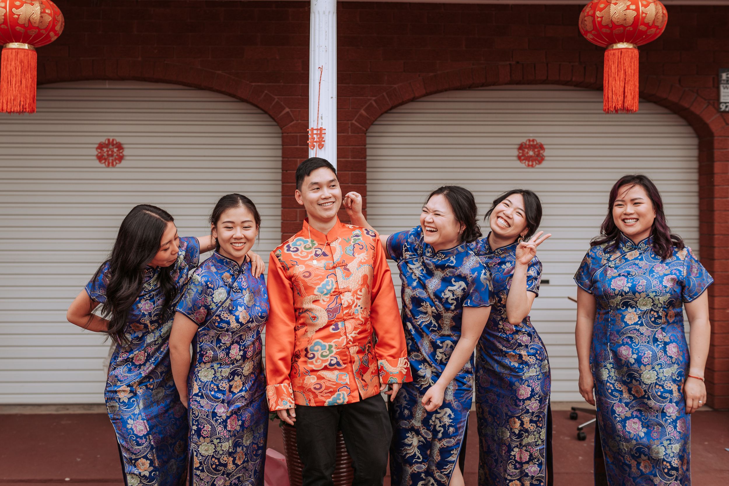 Wedding group photo with bridesmaids in blue qipao dresses and central figure in orange traditional attire.