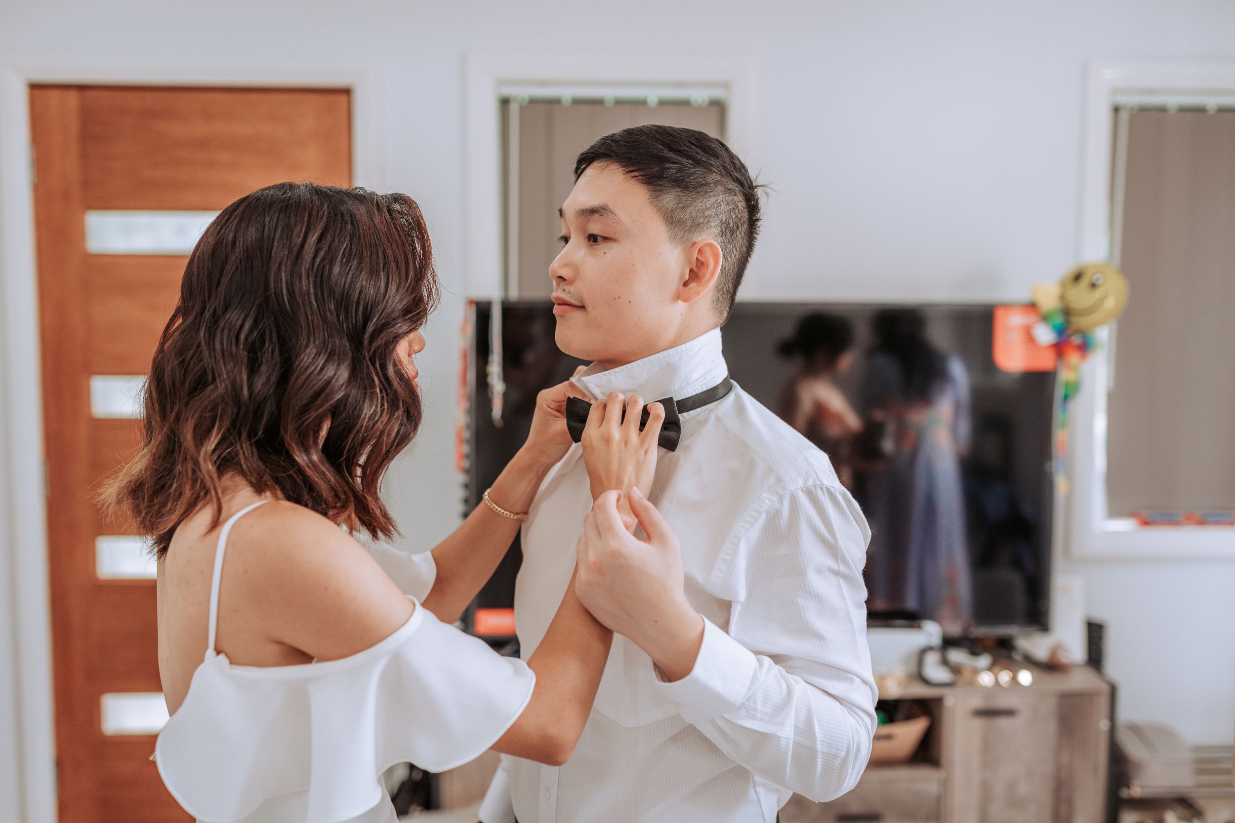 A couple shares an intimate moment in a white off-shoulder dress and formal attire during wedding preparations.