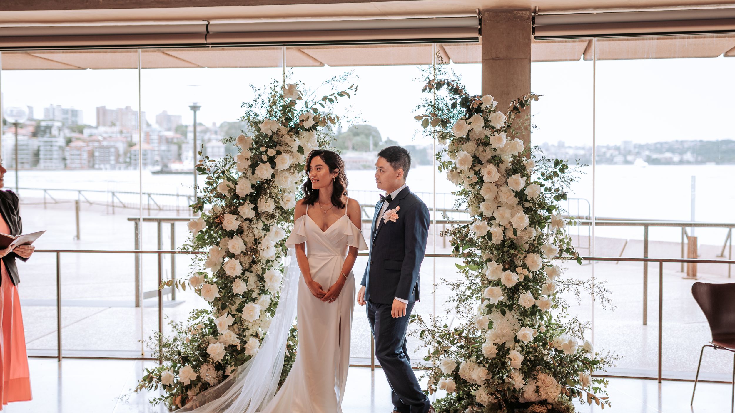 Wedding ceremony under floral arch with pink and white roses against waterfront view.