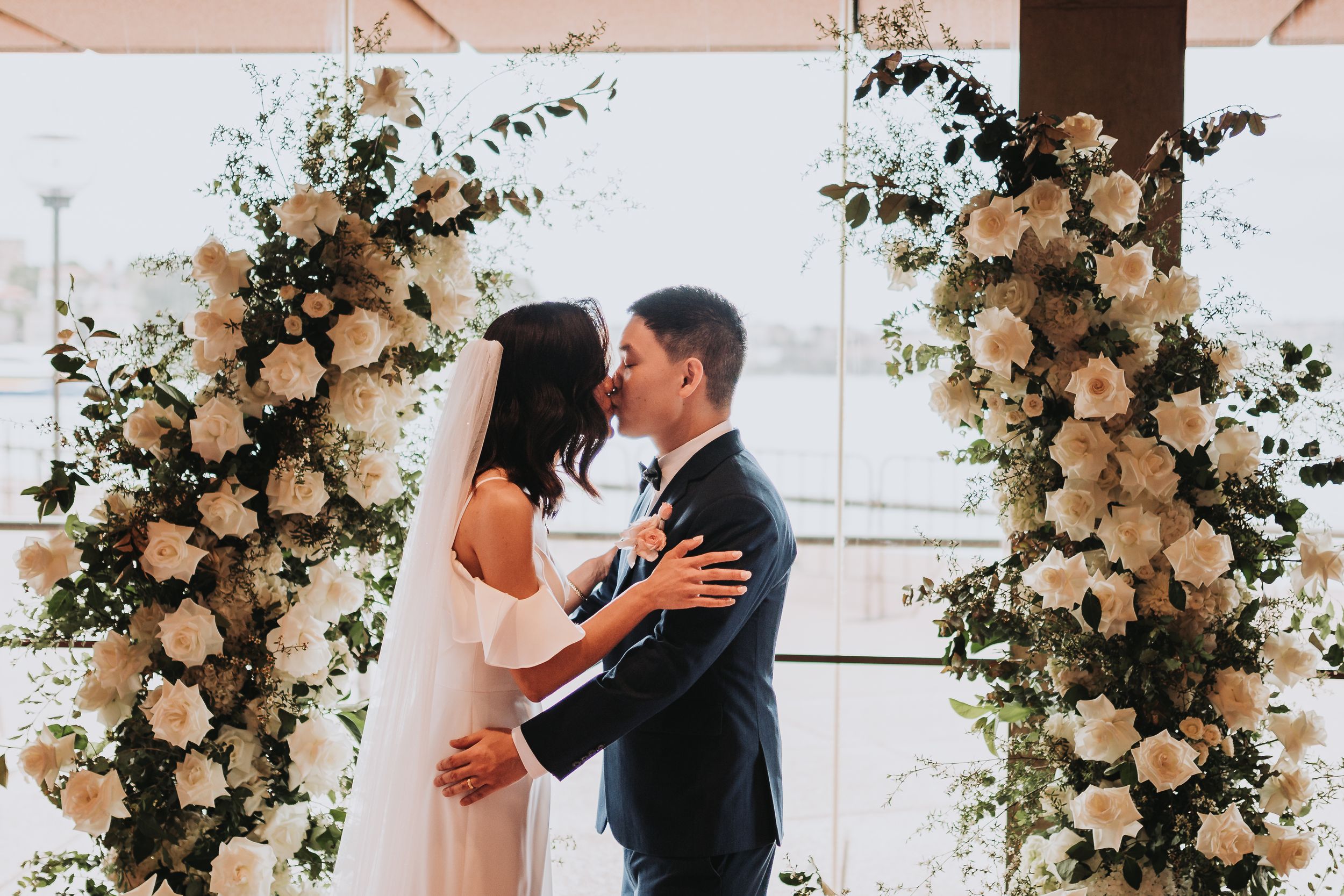 Romantic kiss under white floral arch during wedding ceremony.