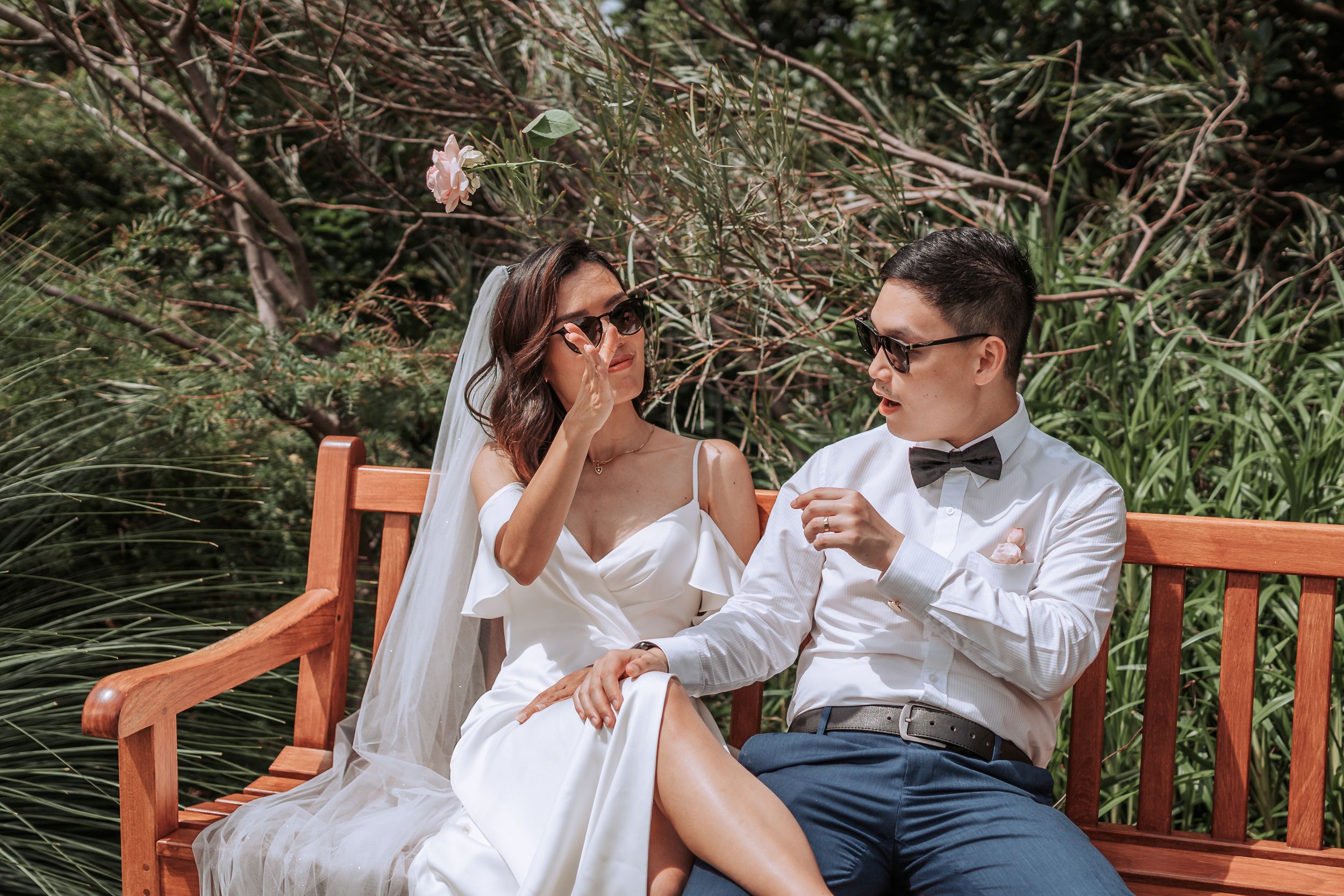 Casual wedding photo shoot on wooden bench surrounded by natural foliage.