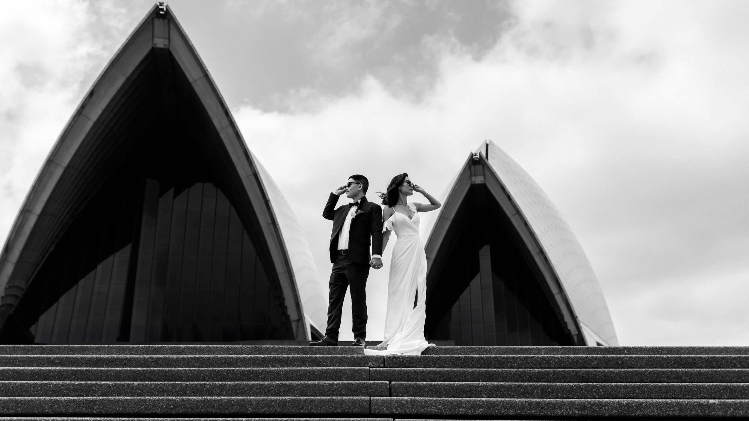A couple in wedding attire stands dramatically against the Sydney Opera House in black and white.