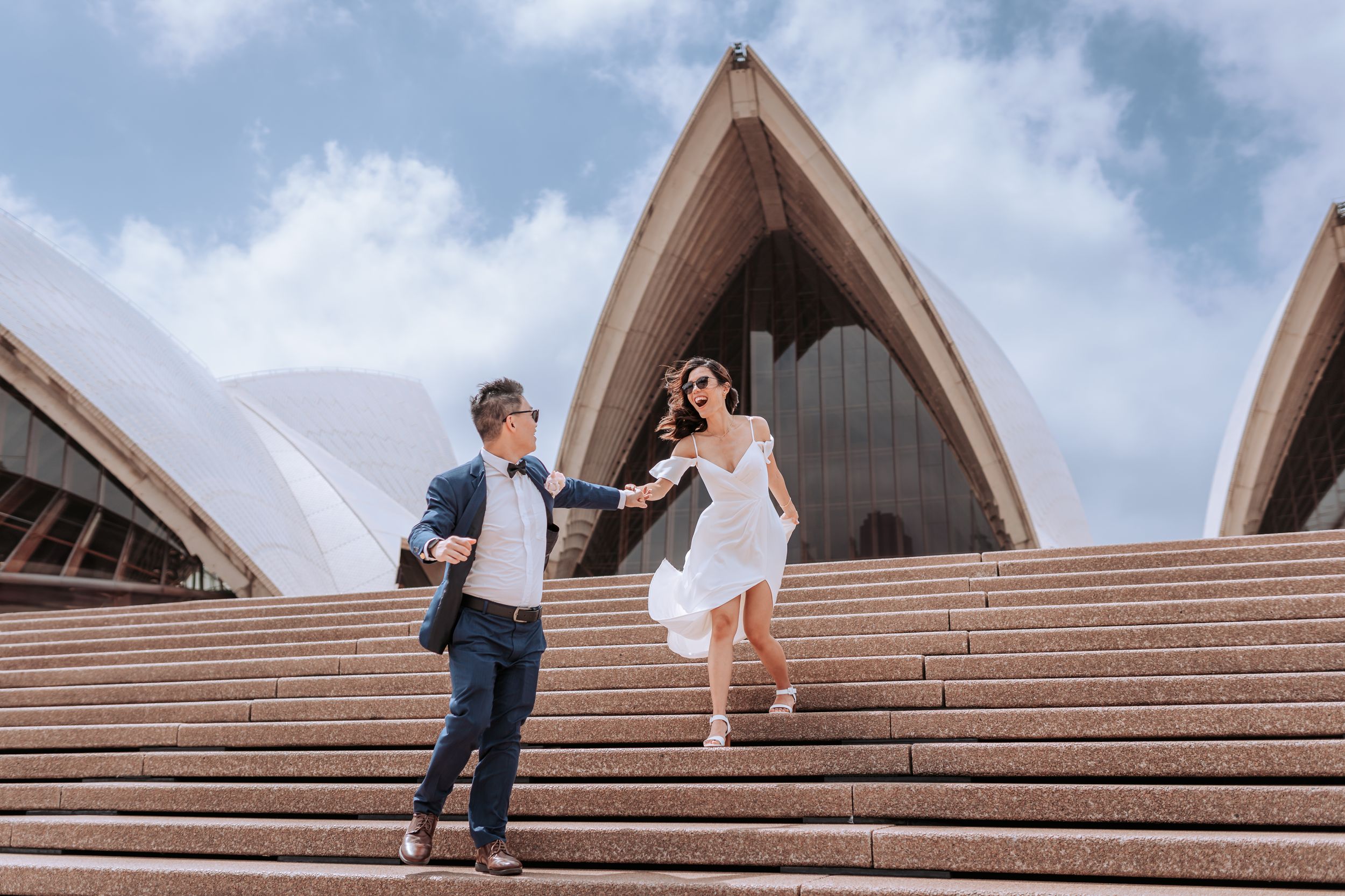 A couple in wedding attire poses playfully on the iconic Sydney Opera House steps.