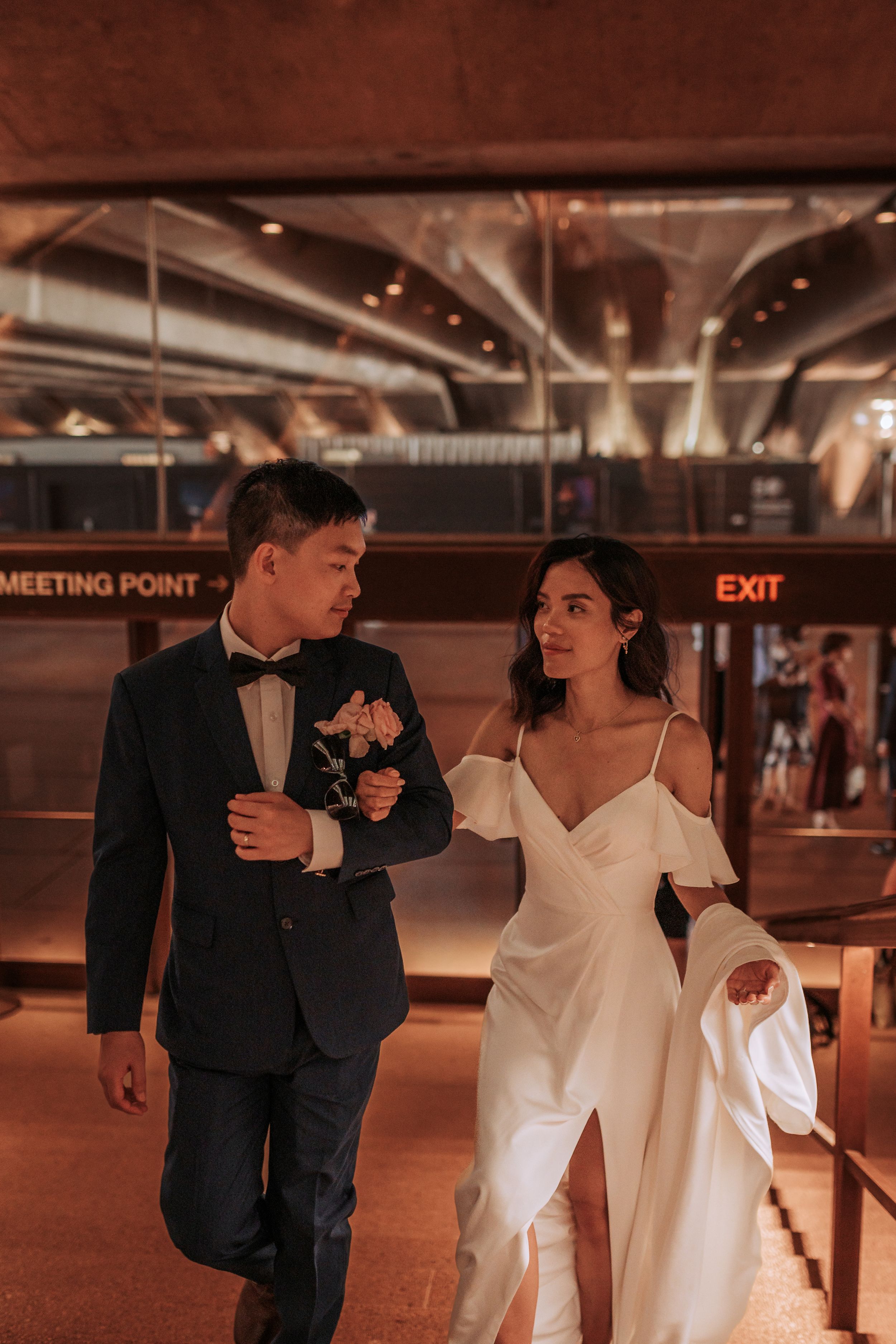 A couple in formal wear walks through an elegantly lit indoor venue.