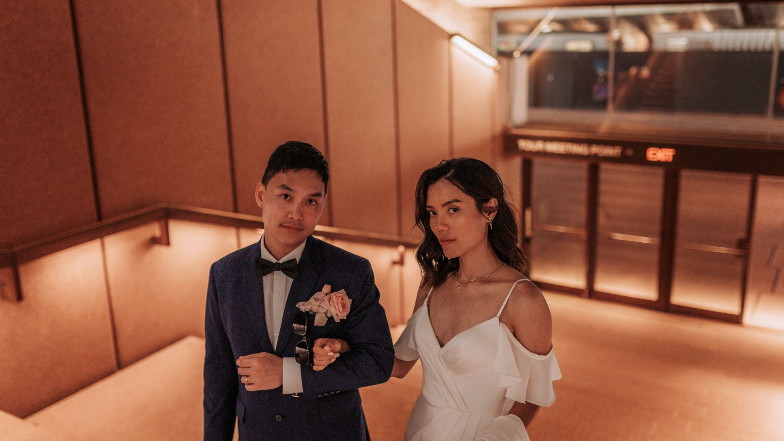 A couple in formal attire strolls through a warmly lit indoor corridor.