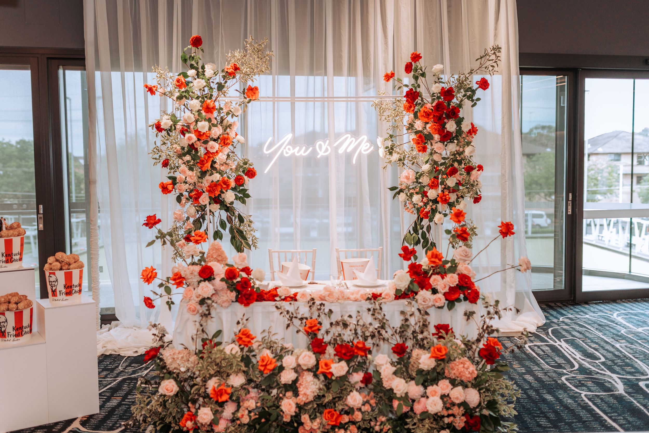 Elegant wedding backdrop with red and pink floral arrangements framing a neon sign against sheer white curtains.