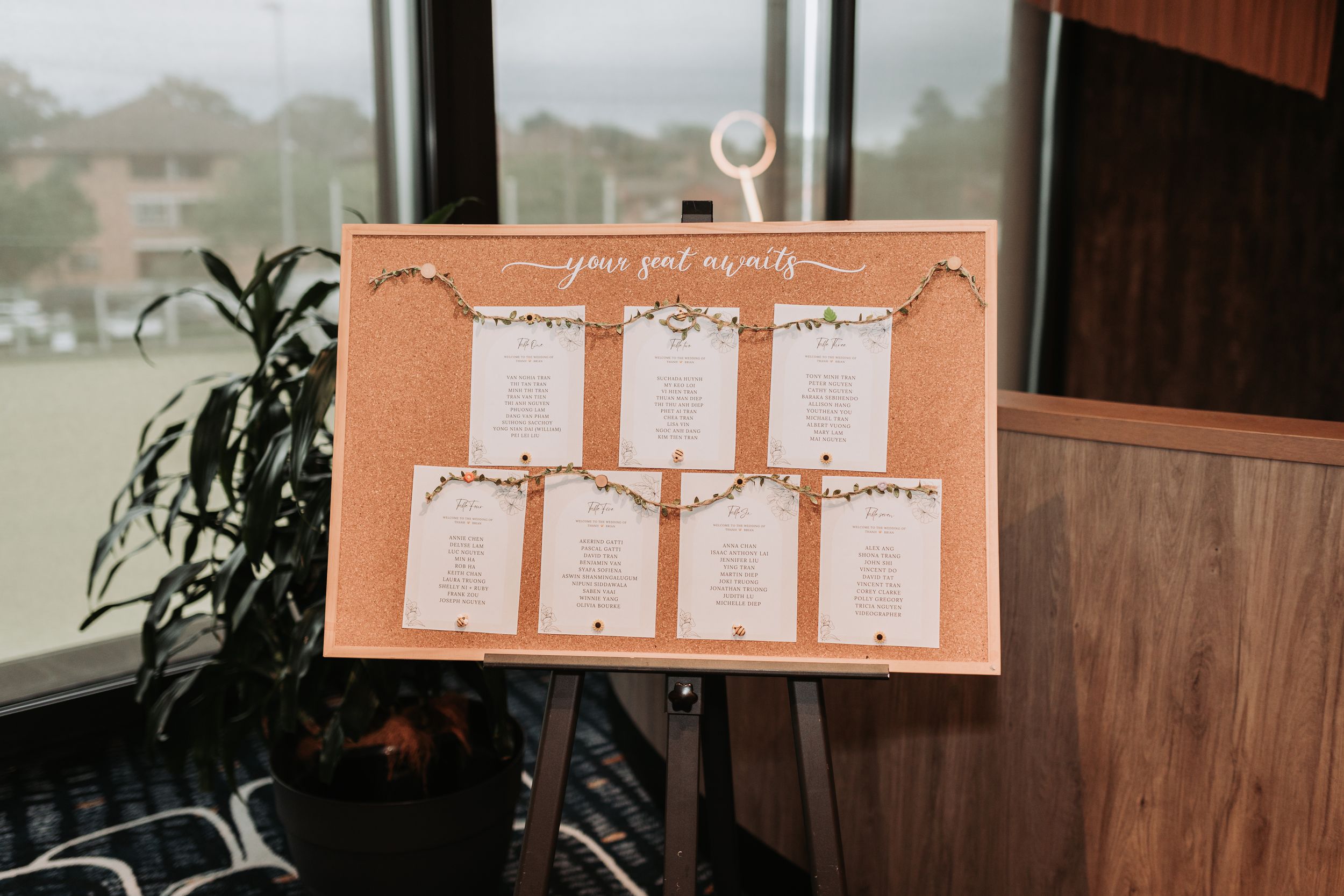 Peach-colored wedding seating chart display board mounted on an easel near a window.