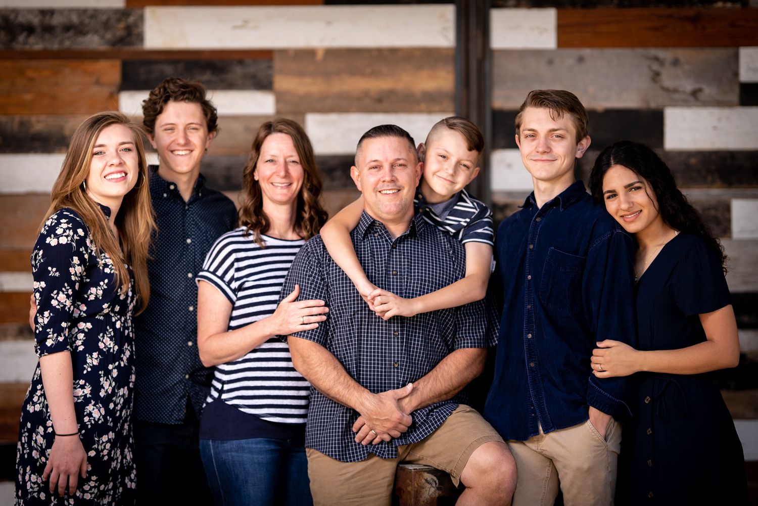 A group of seven people dressed in casual attire posing for a portrait against a rustic wooden wall background.
