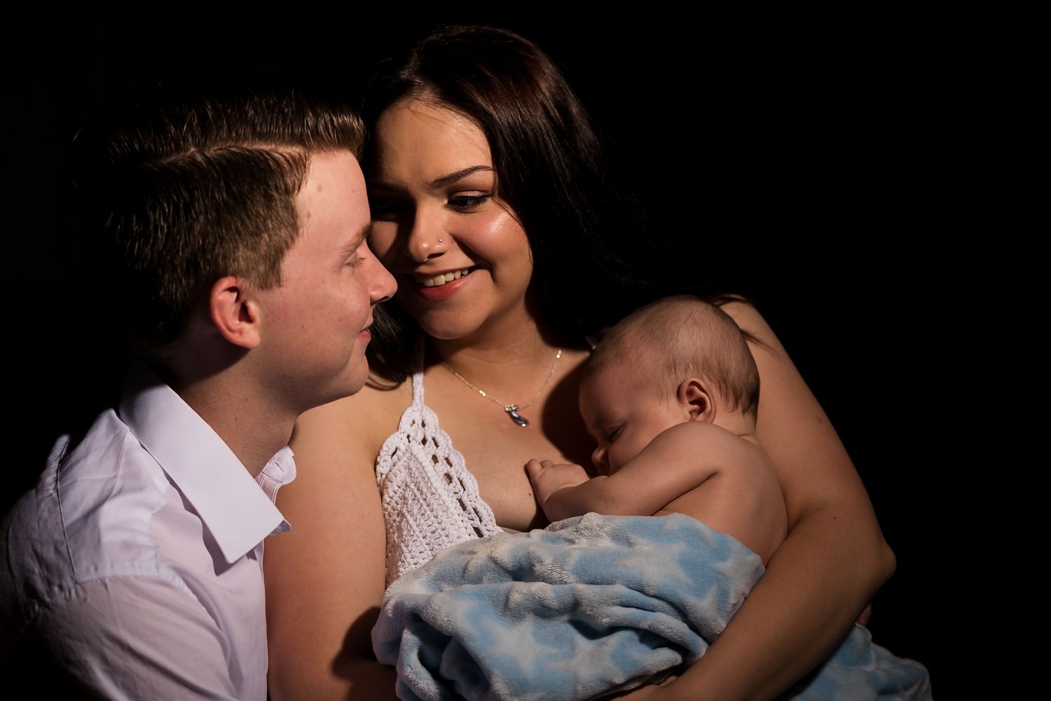 A family sharing an intimate moment together with a baby wrapped in a blue blanket in soft dramatic lighting.