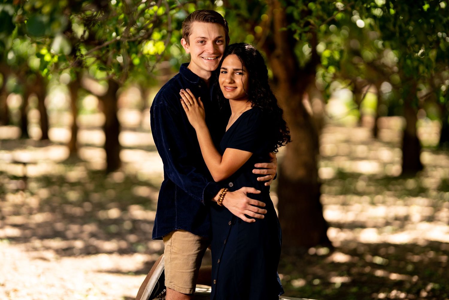 A romantic couple embrace in a sun-dappled orchard wearing coordinating navy blue outfits.