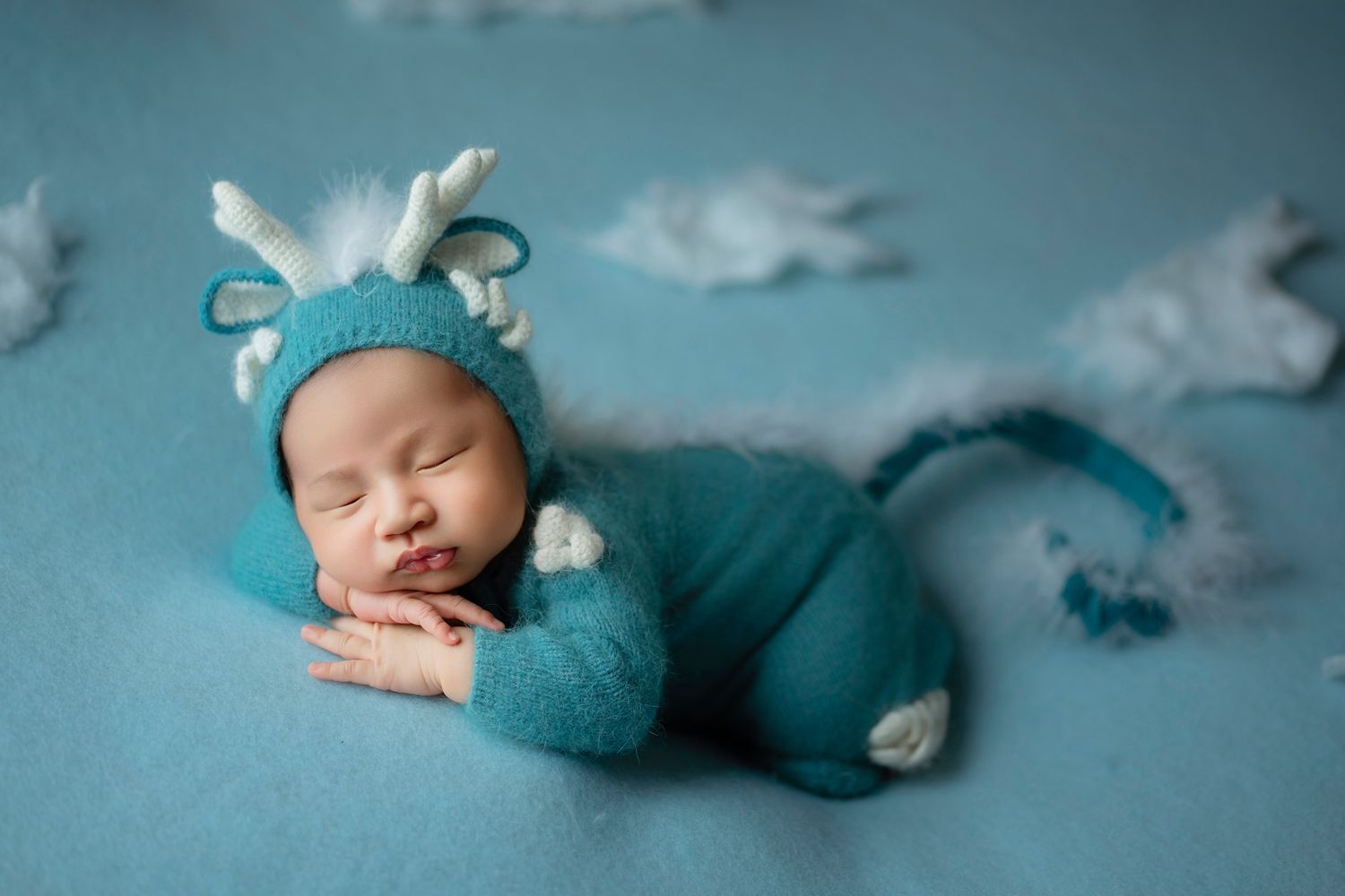 Sleeping newborn wearing teal knit animal outfit on blue backdrop with cotton ball clouds.