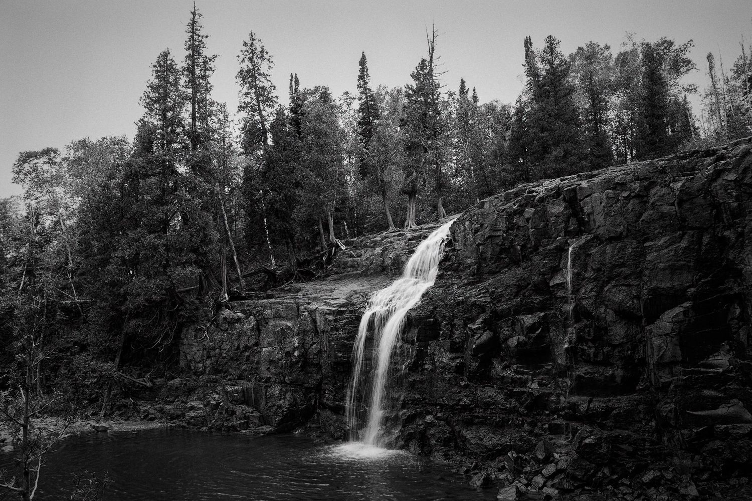 A black and white scenic waterfall cascades down rocky cliffs surrounded by evergreen trees in a forested landscape.