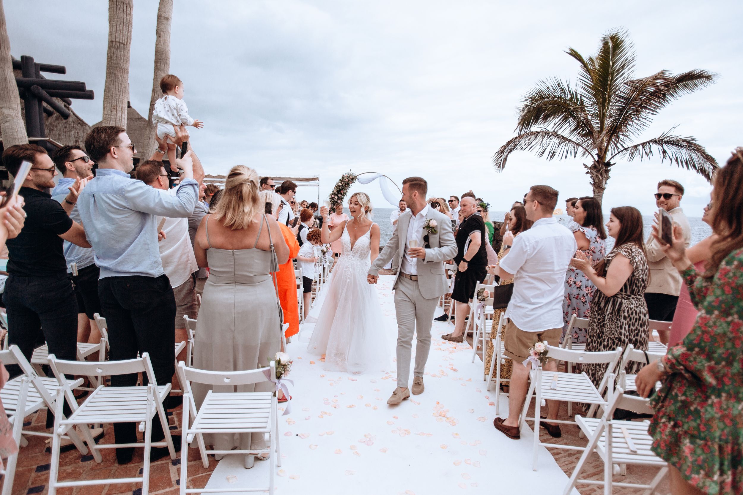 Beach wedding ceremony with guests standing and celebrating as couple walks down aisle.