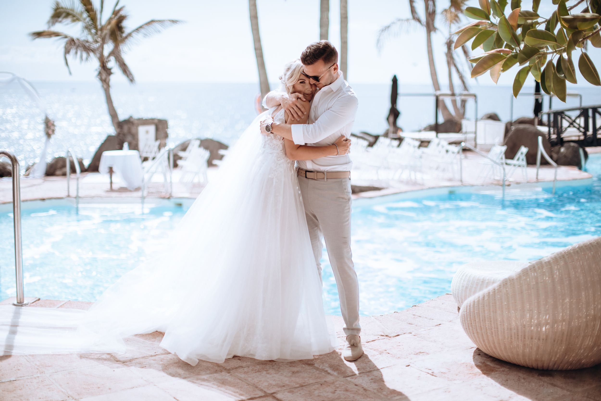 Wedding couple poses by poolside at luxurious tropical resort with palm trees.