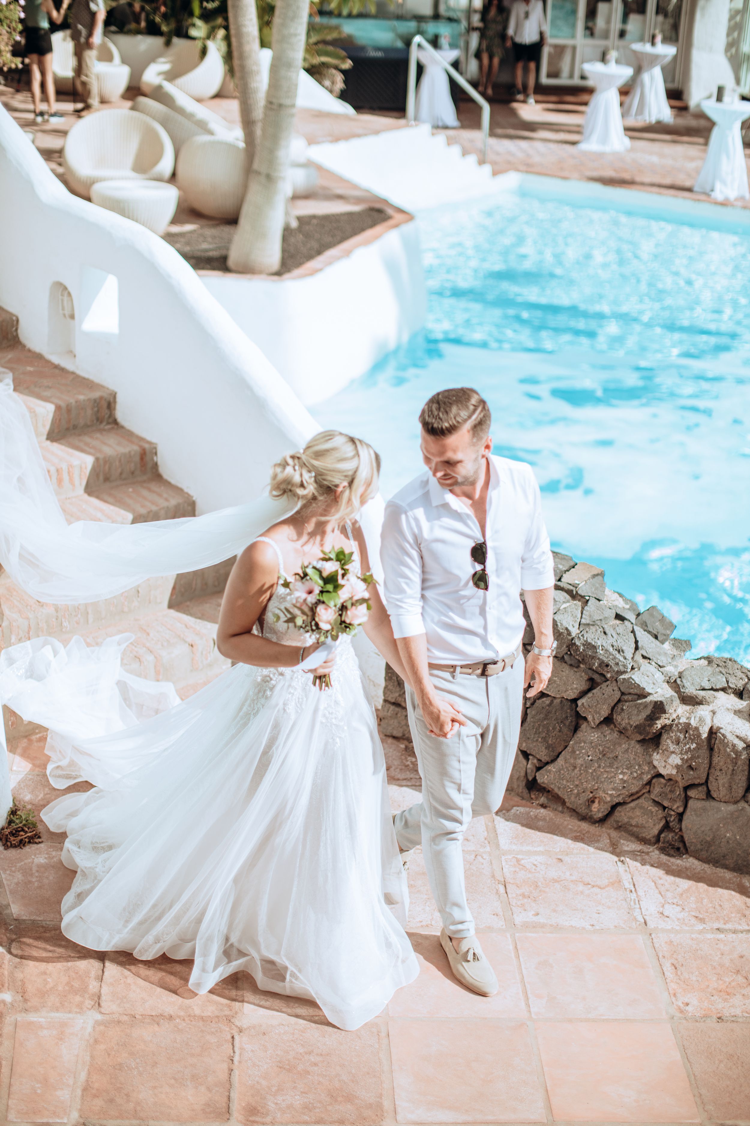 Wedding couple walking along poolside pathway at tropical resort with flowing white dress.