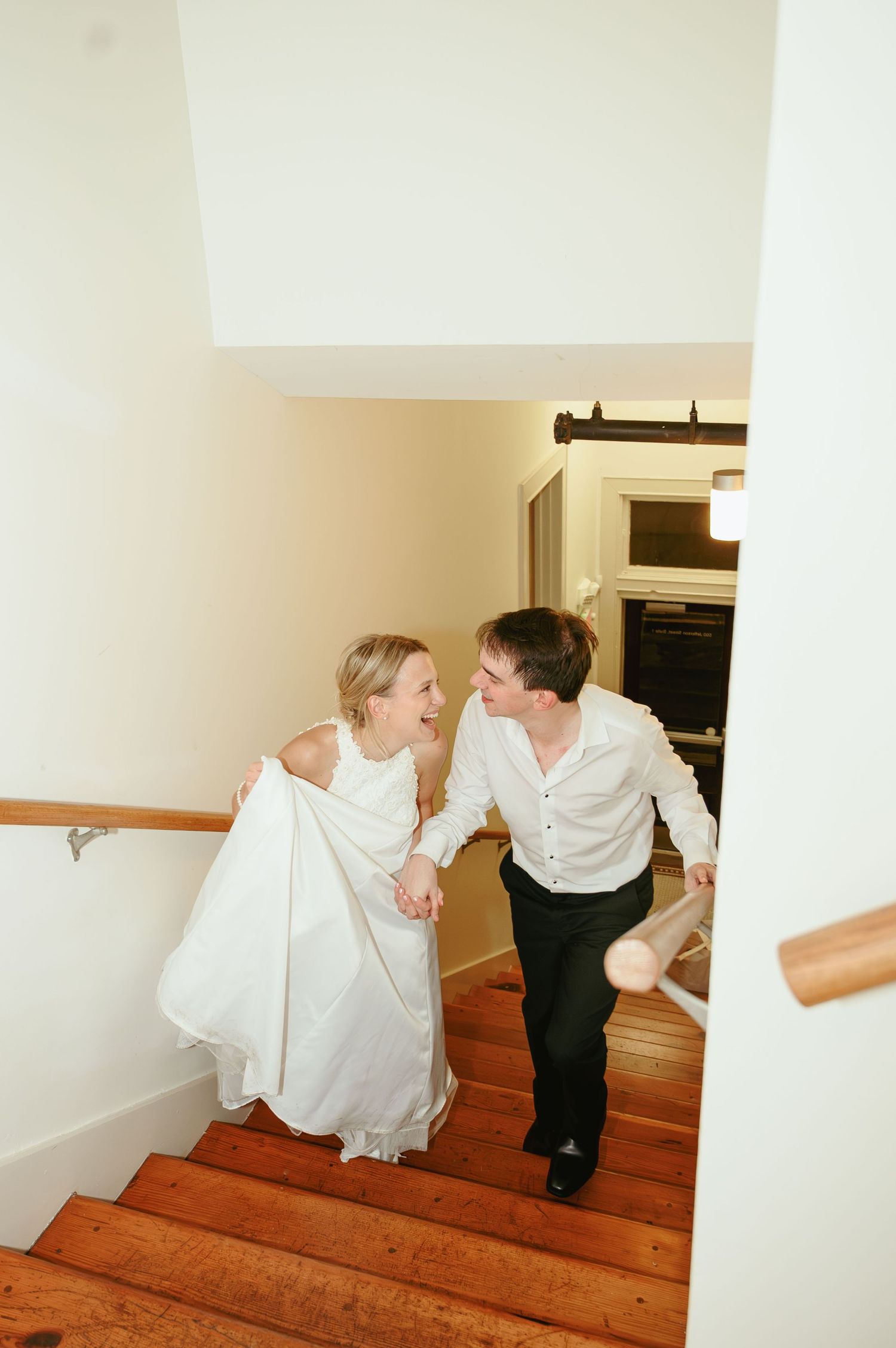 Two people in formal attire laugh together while climbing wooden stairs in an indoor setting.