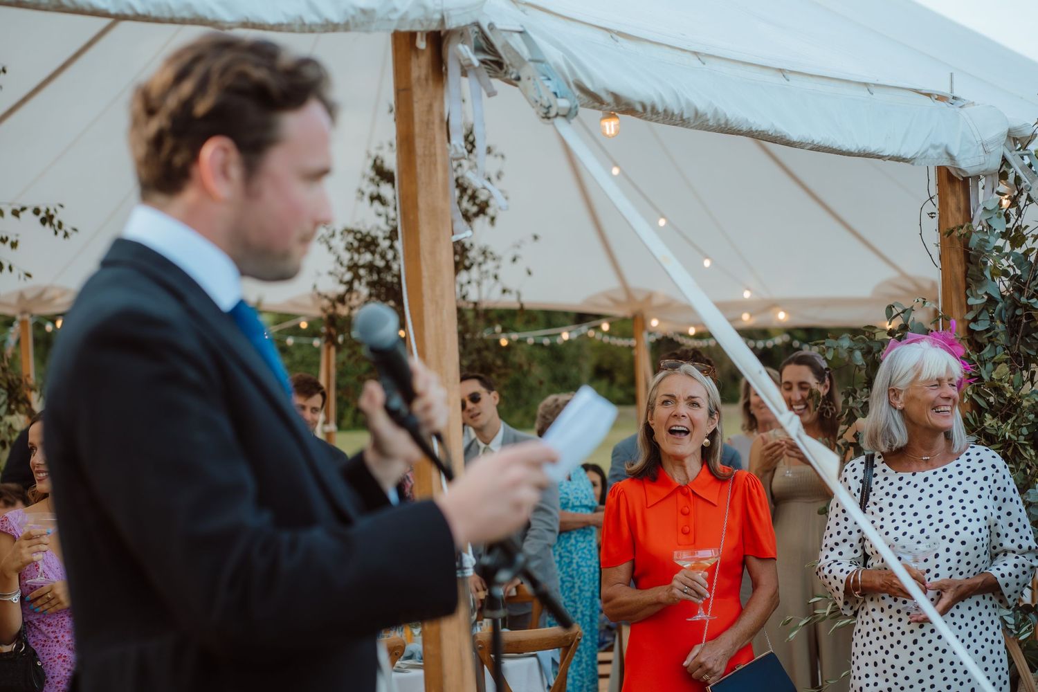 Guests at a wedding reception raise their glasses in celebration during an outdoor toast at a tent venue.