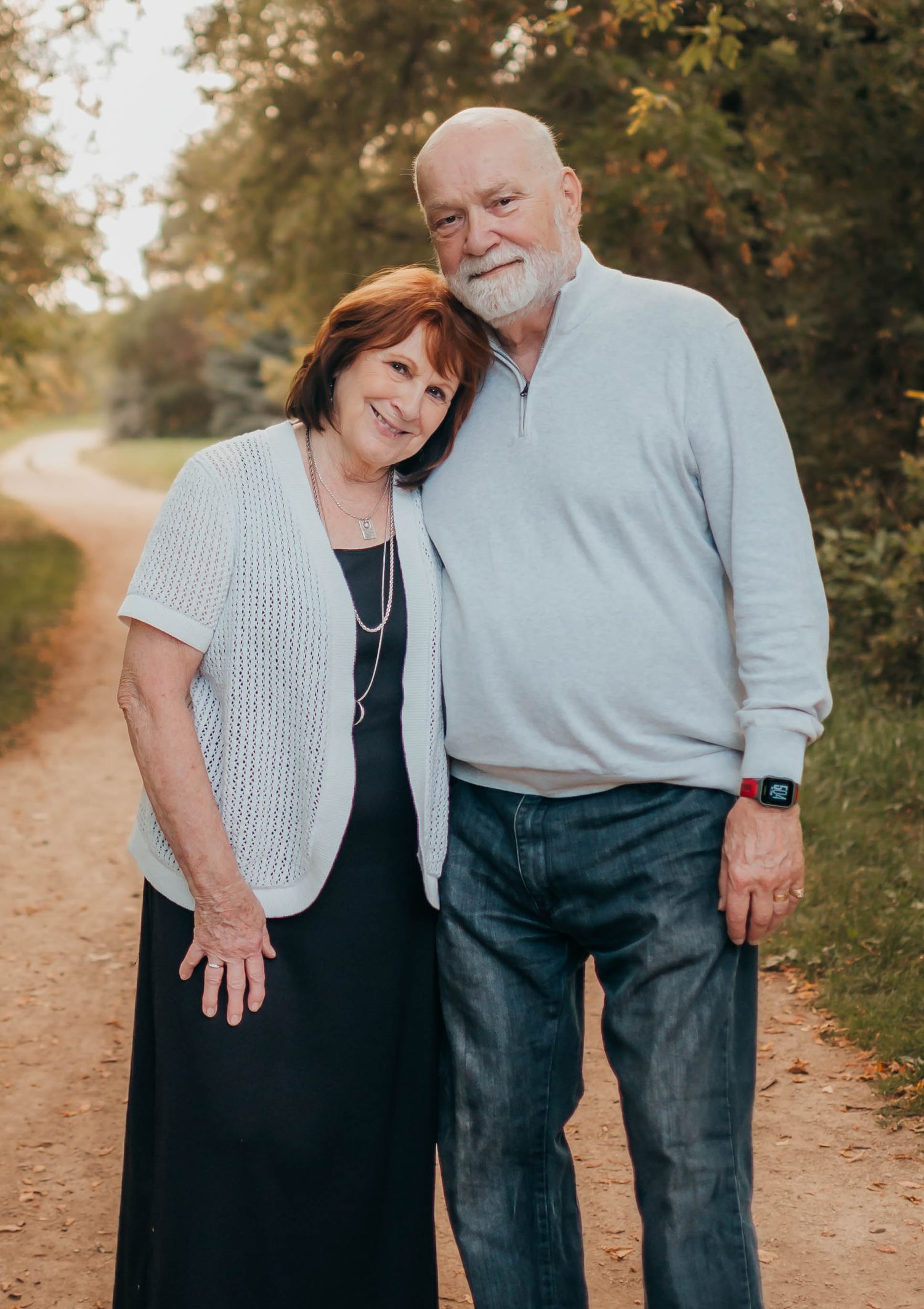Smiling mature couple stands together on a dirt path during golden hour with autumn trees in the background.