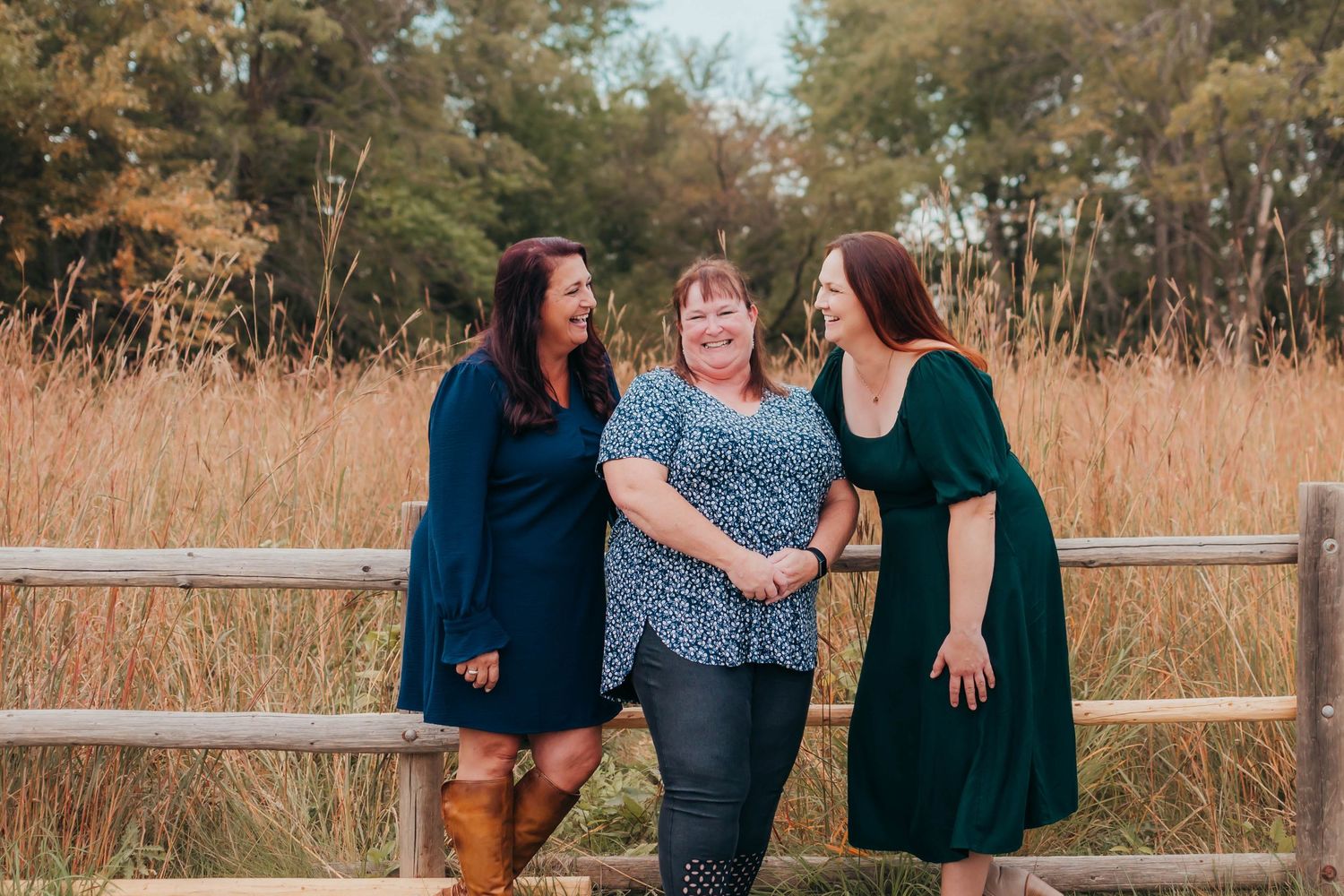 Three people in blue and green dresses share a candid moment laughing together by a wooden fence in a rustic field setting.