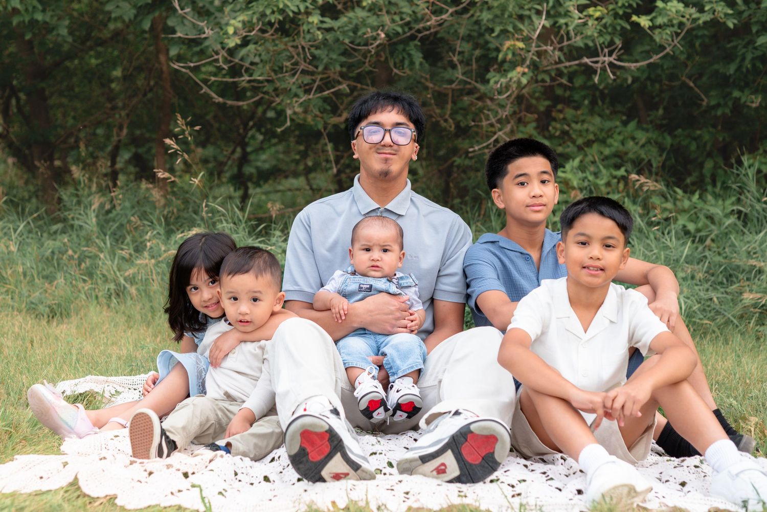 A group in casual attire sits together on a white blanket outdoors with greenery in the background.