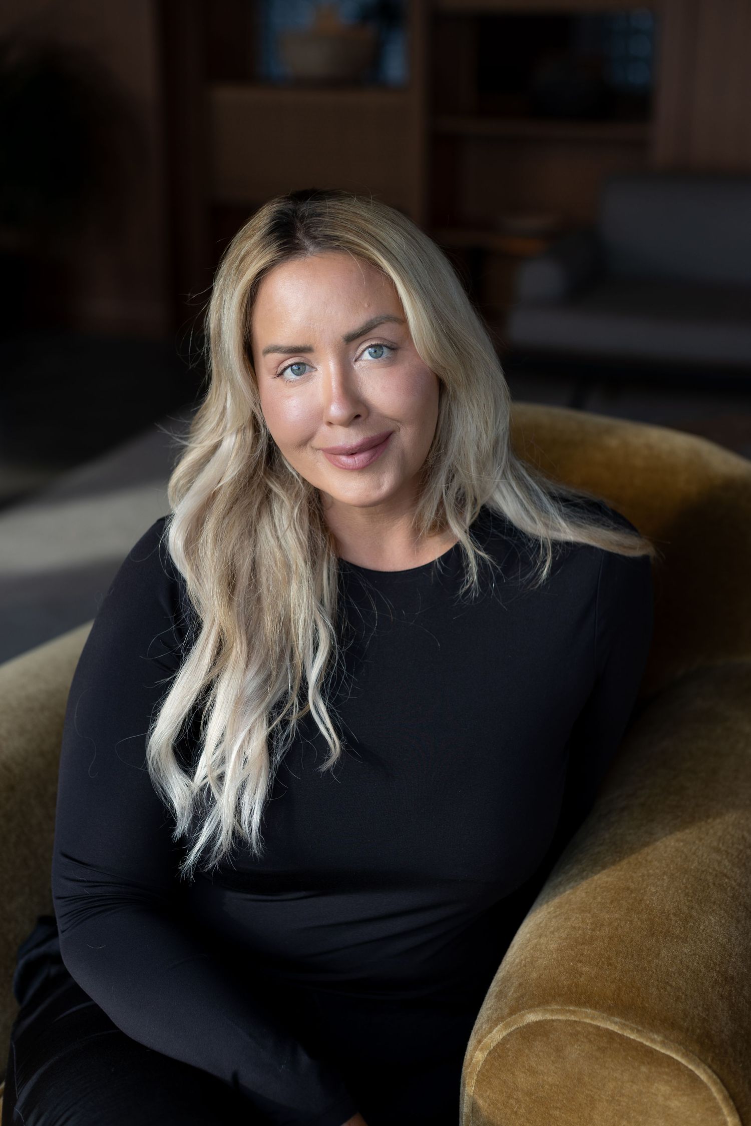 Portrait of someone with long blonde hair wearing a black long-sleeve dress seated casually in an indoor setting.
