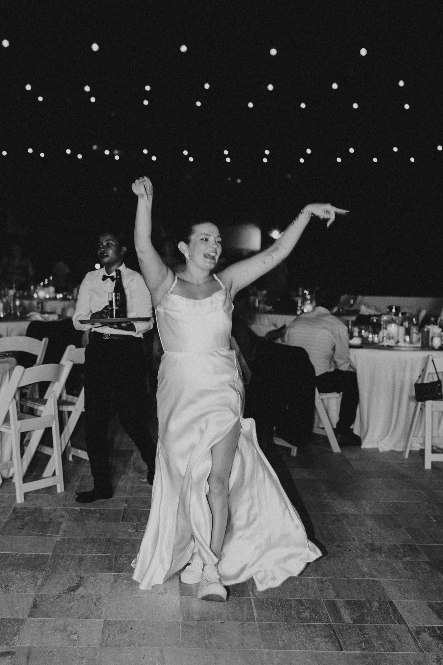 A person in a wedding dress dances joyfully under string lights at an outdoor reception.