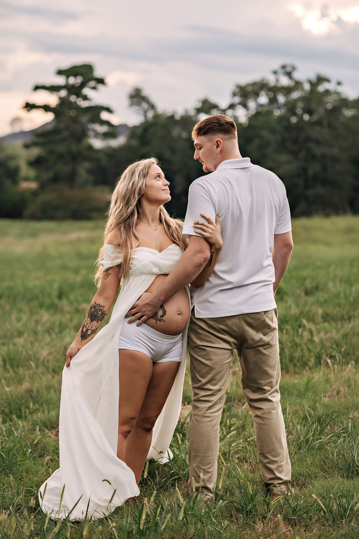 Romantic couple embraces in a scenic meadow at sunset, with tall trees and mountains in the background.