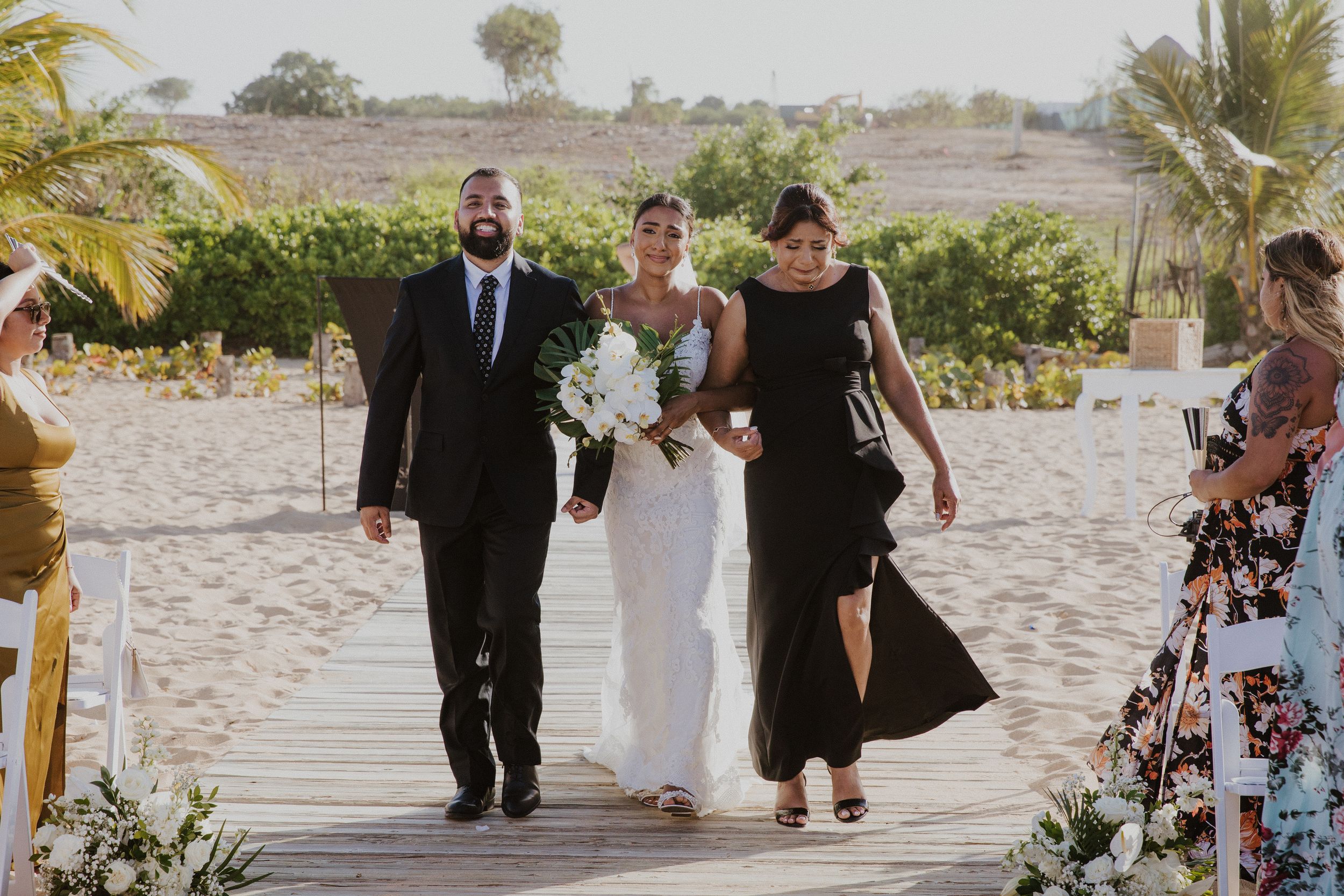 Wedding ceremony procession on beach with palm trees and guests in formal attire.