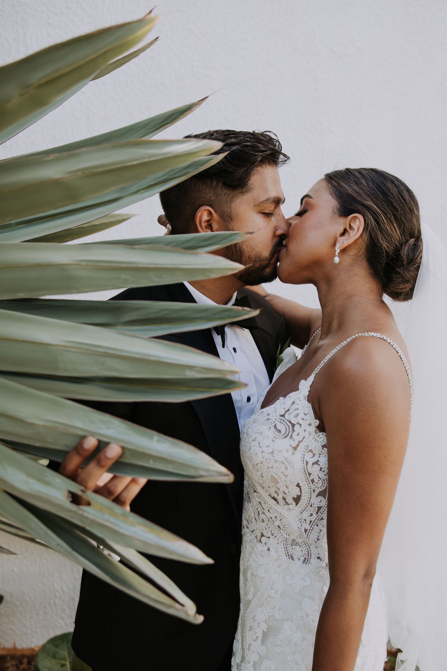 Romantic wedding couple embracing near tropical palm leaves against white architectural backdrop.
