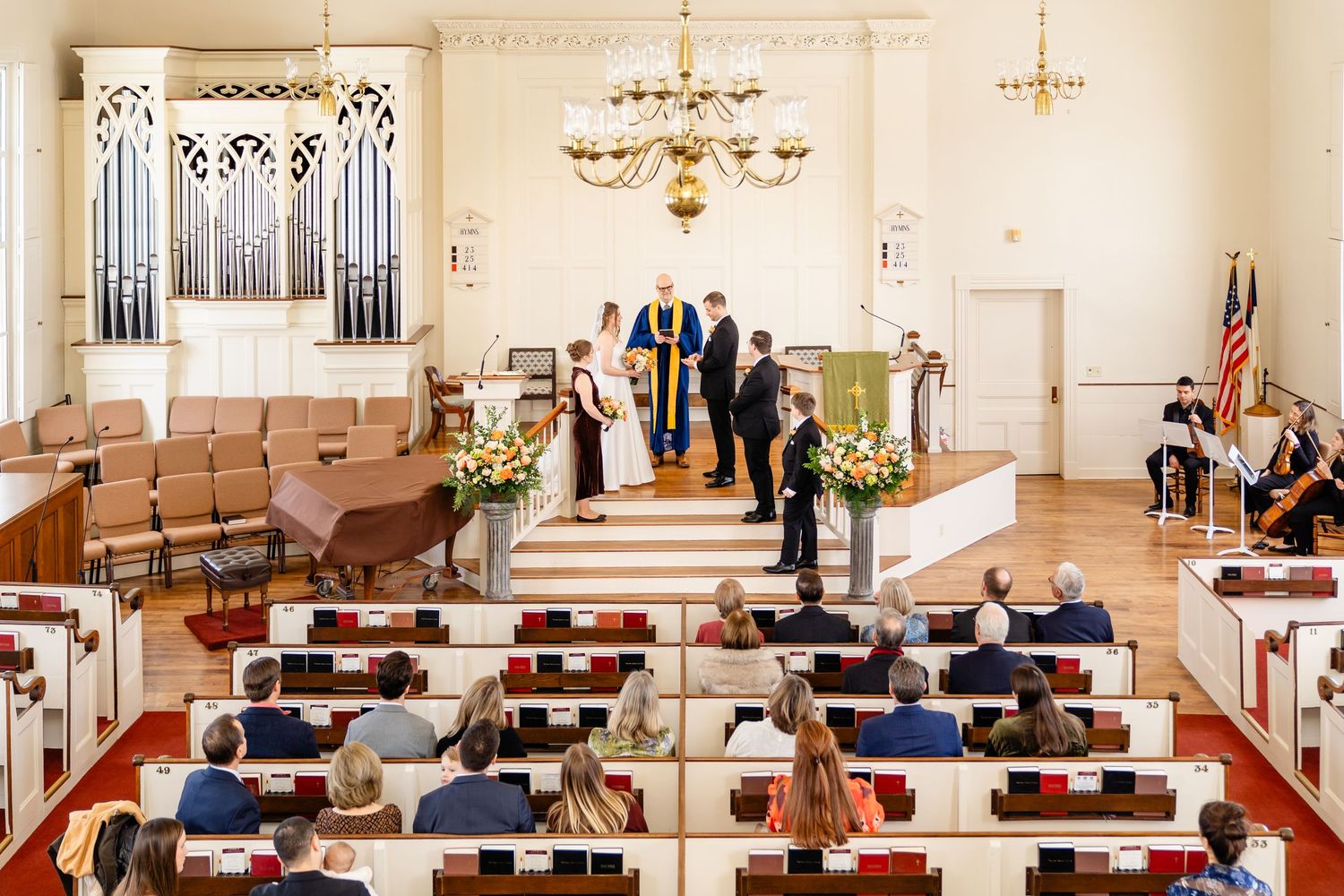 People are gathered for a traditional church wedding ceremony in a bright, elegant chapel with wooden pews and chandeliers.