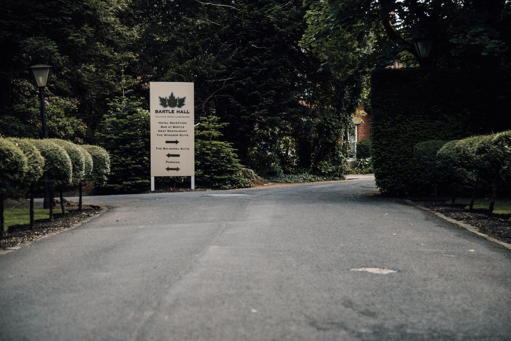 Tree-lined driveway entrance to a venue with a rustic wooden sign at the end of the path.