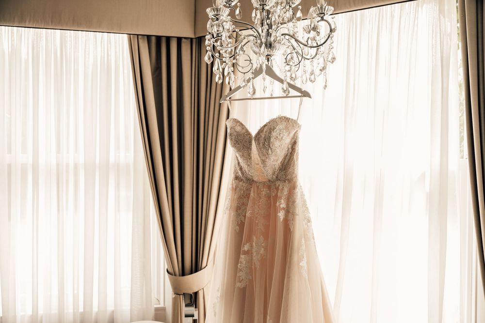 Elegant wedding dress hanging in a luxurious room with chandelier and draped windows.