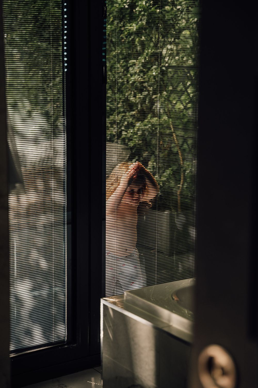 Sunlight streams through venetian blinds onto a figure standing by a window.