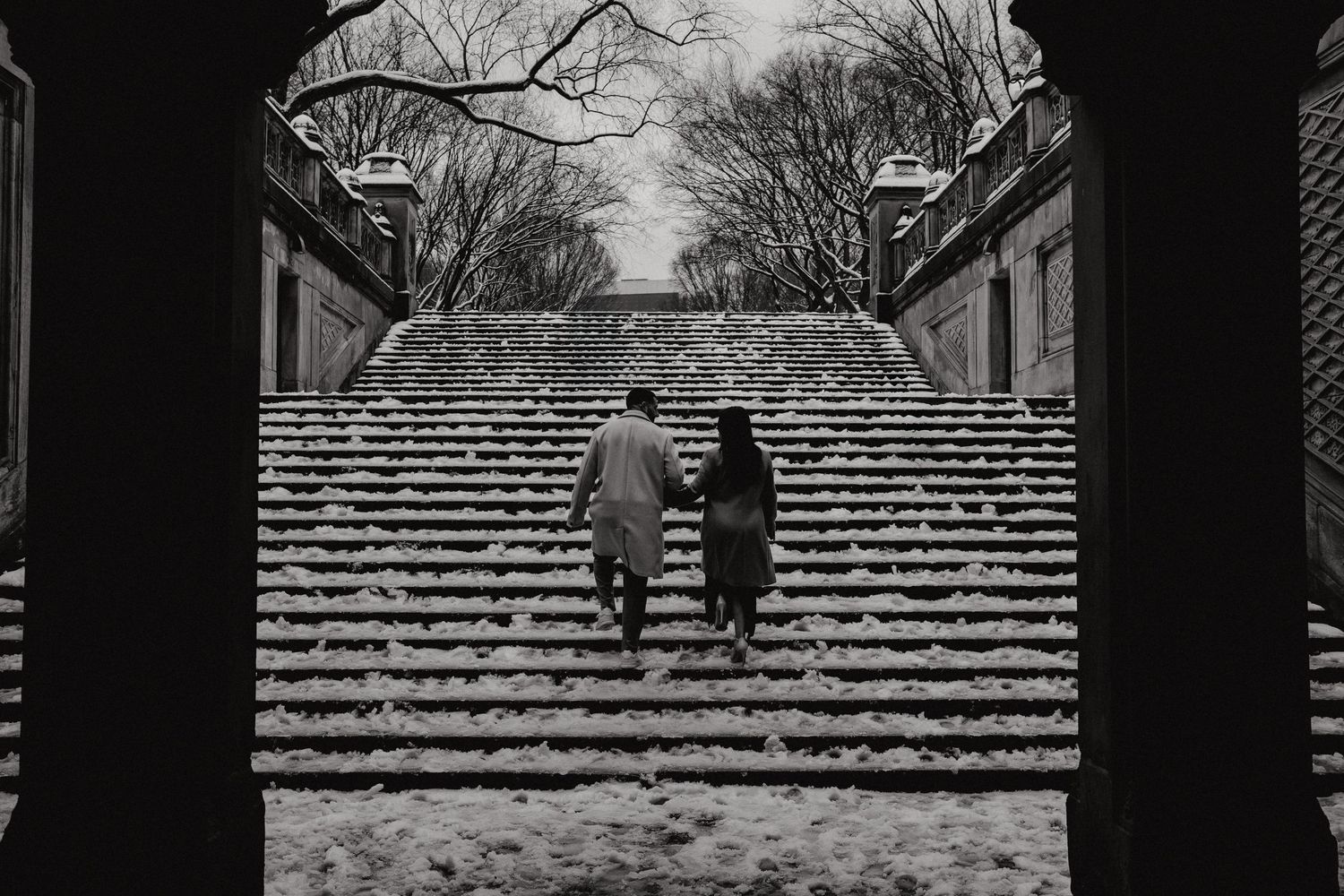 A couple ascending snow-covered stairs in Central Park framed by an architectural arch in black and white.