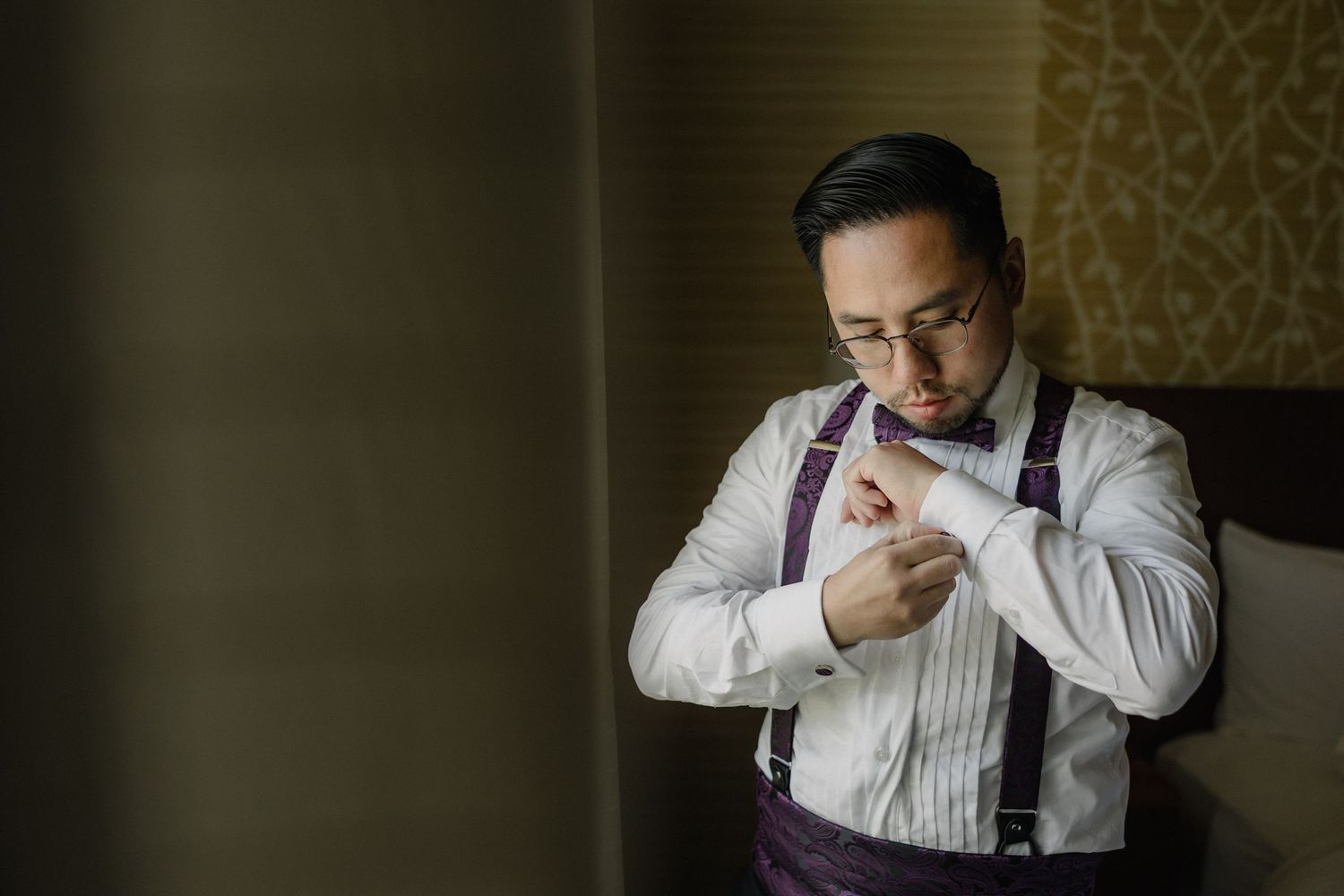 A person in suspenders and white dress shirt adjusts their cuff by a window in moody lighting.