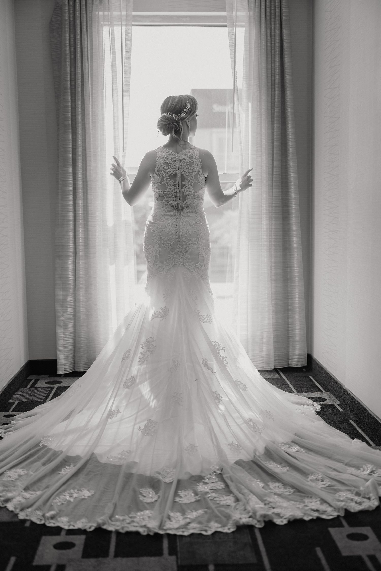 A bride in a stunning lace mermaid wedding gown stands silhouetted against a sunlit window with flowing train behind her.