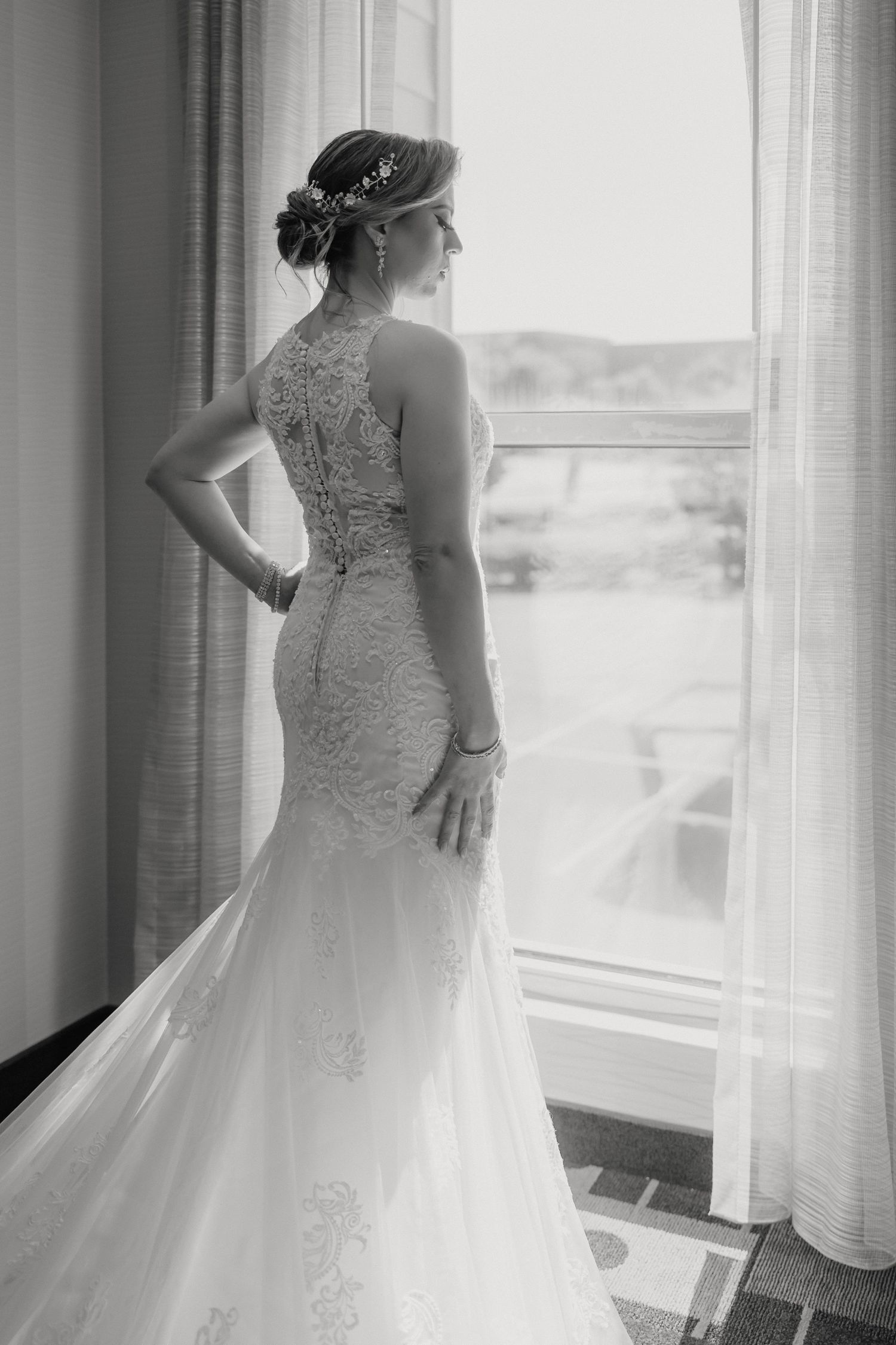 A bride in an elegant lace-detailed mermaid wedding gown gazes out a window in black and white photography.