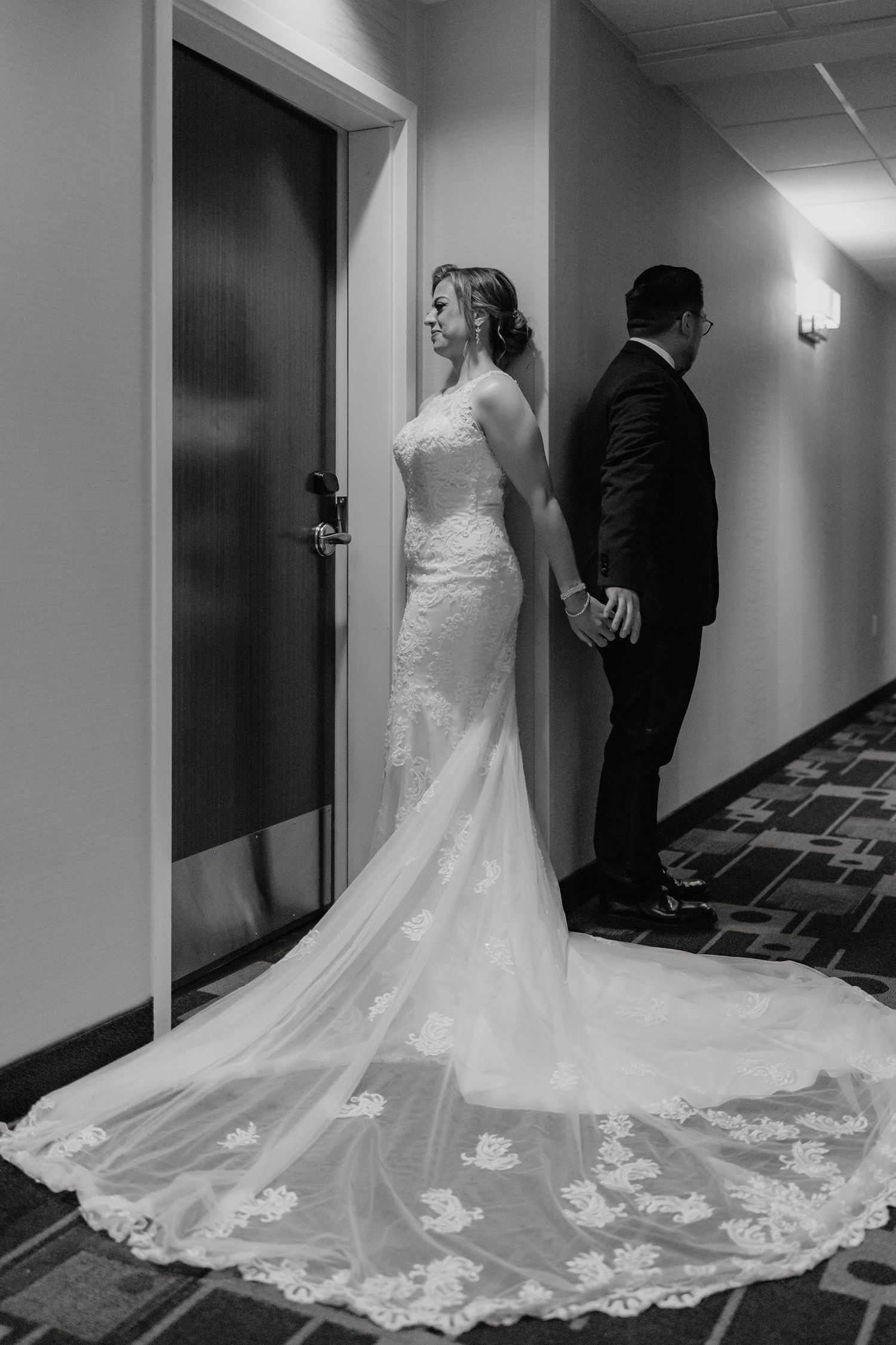 A couple shares a private first look moment in a hotel hallway, with a stunning lace train wedding gown on display.