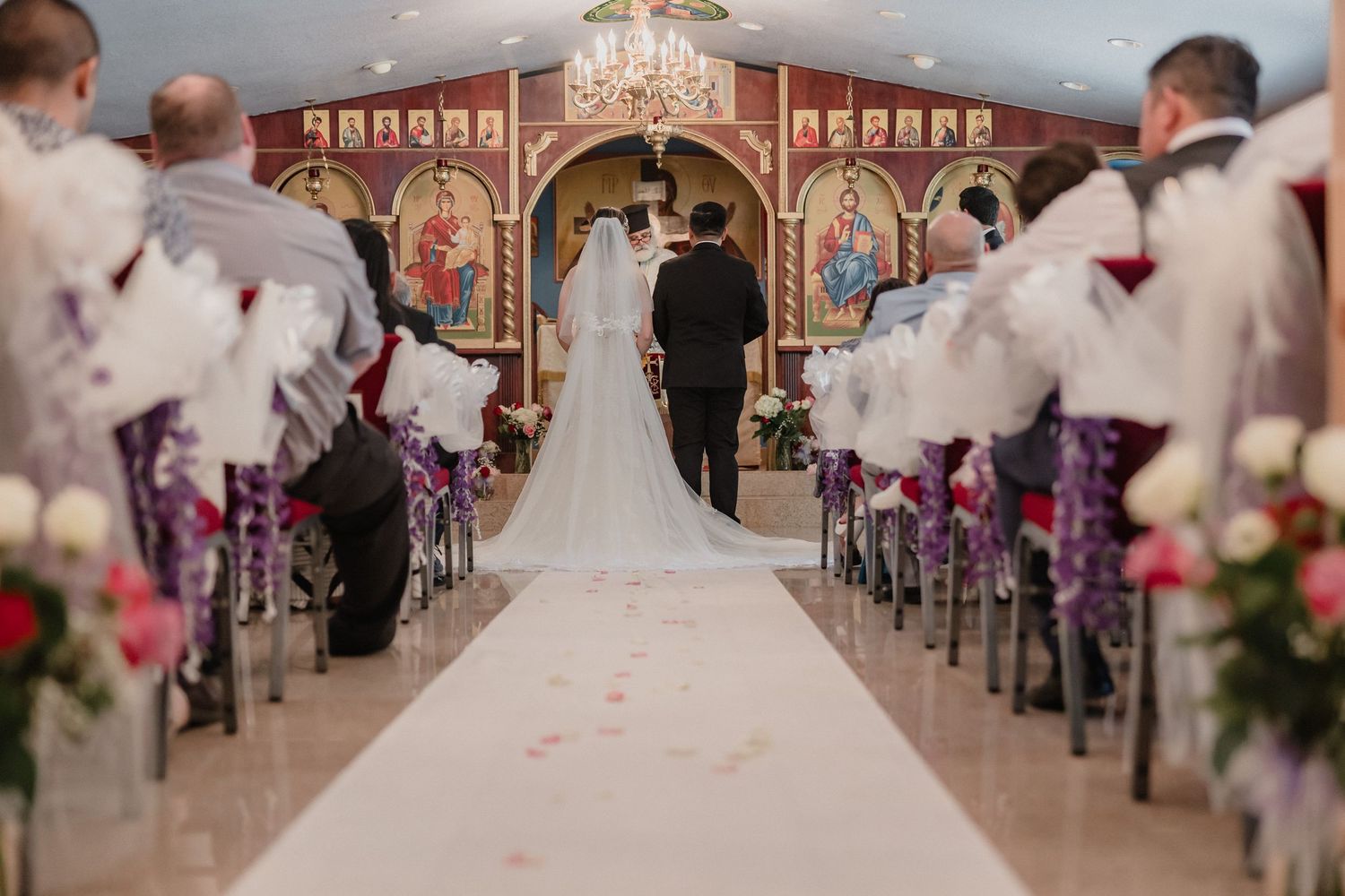 Traditional wedding ceremony in a decorated church with rose petals lining the aisle and elegant chandelier lighting.