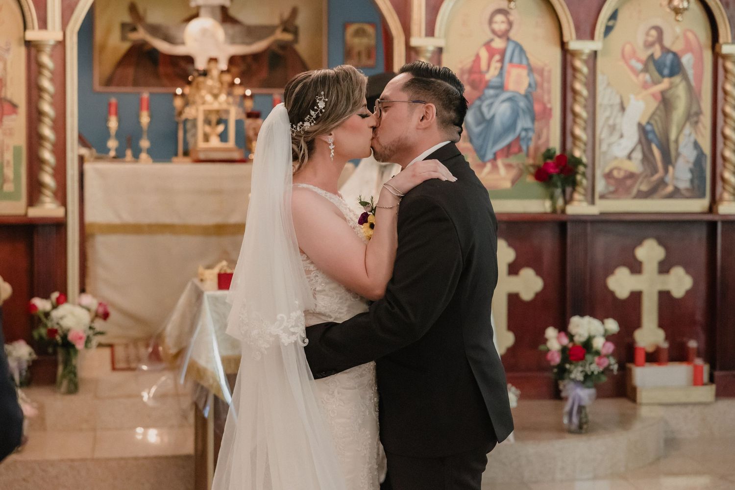 A tender wedding moment between a couple sharing a kiss at the altar inside an ornate church with religious iconography.