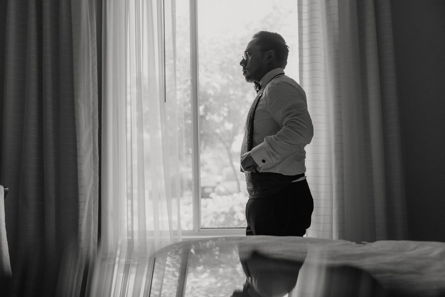A silhouetted figure in business attire gazes through a window with flowing curtains in black and white.
